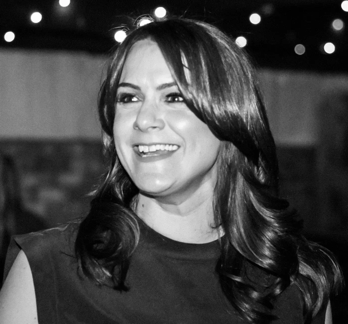 A black and white photo of a woman smiling. She has long, dark hair and is wearing a sleeveless top.