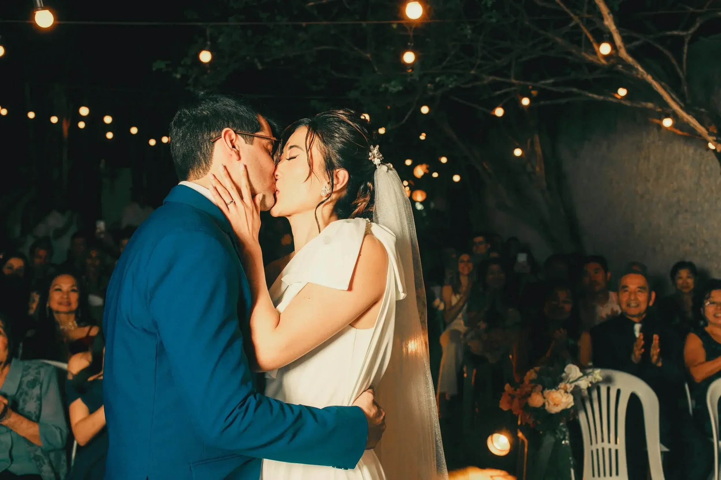 A couple is sharing a kiss during their wedding ceremony, surrounded by guests under fairy lights at night.