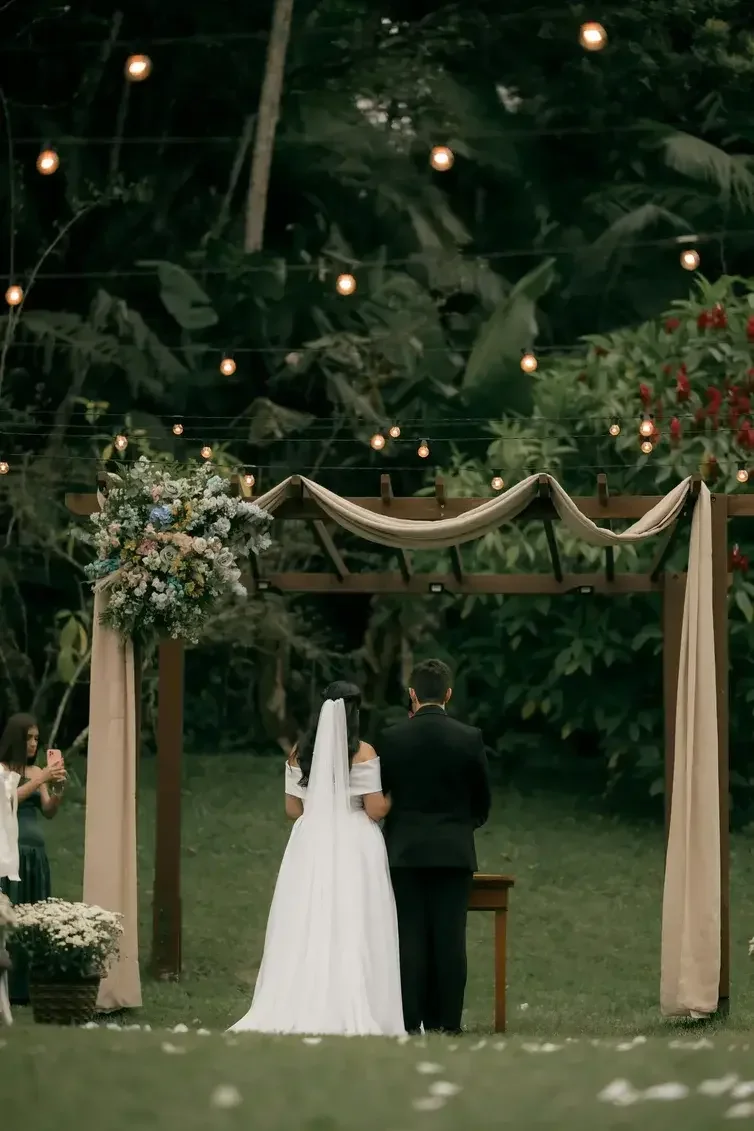 A bride and groom standing under a decorated wooden wedding arch in an outdoor setting with string lights and lush greenery, surrounded by guests.