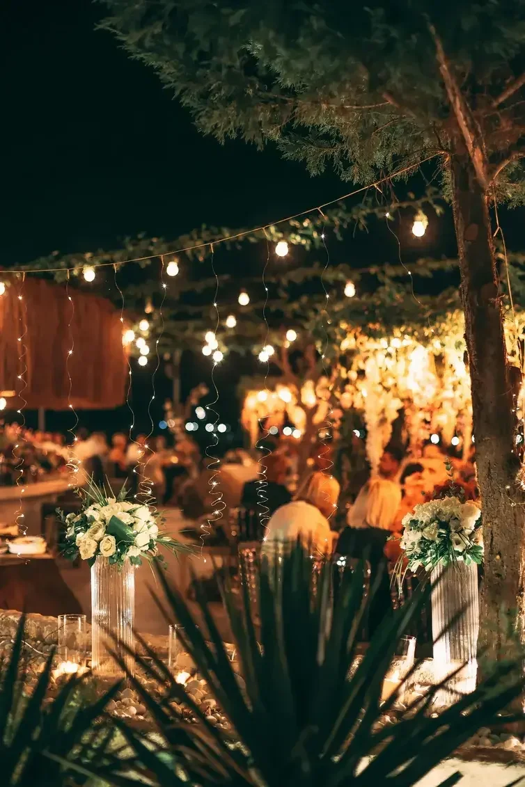 Outdoor evening celebration with string lights, floral arrangements, and people socializing under trees.
