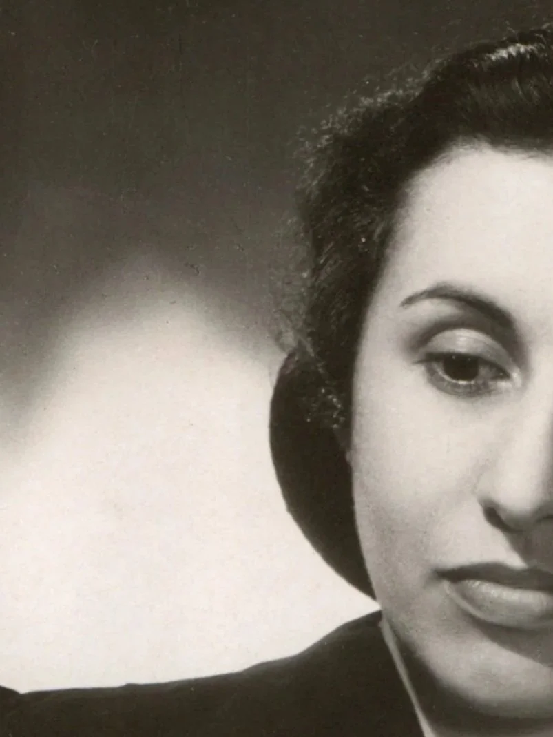A black-and-white photo of a woman with dark hair styled in waves, wearing makeup with defined eyebrows, eyeliner, and lipstick. She is dressed in vintage style, possibly from the 1940s or 1950s.