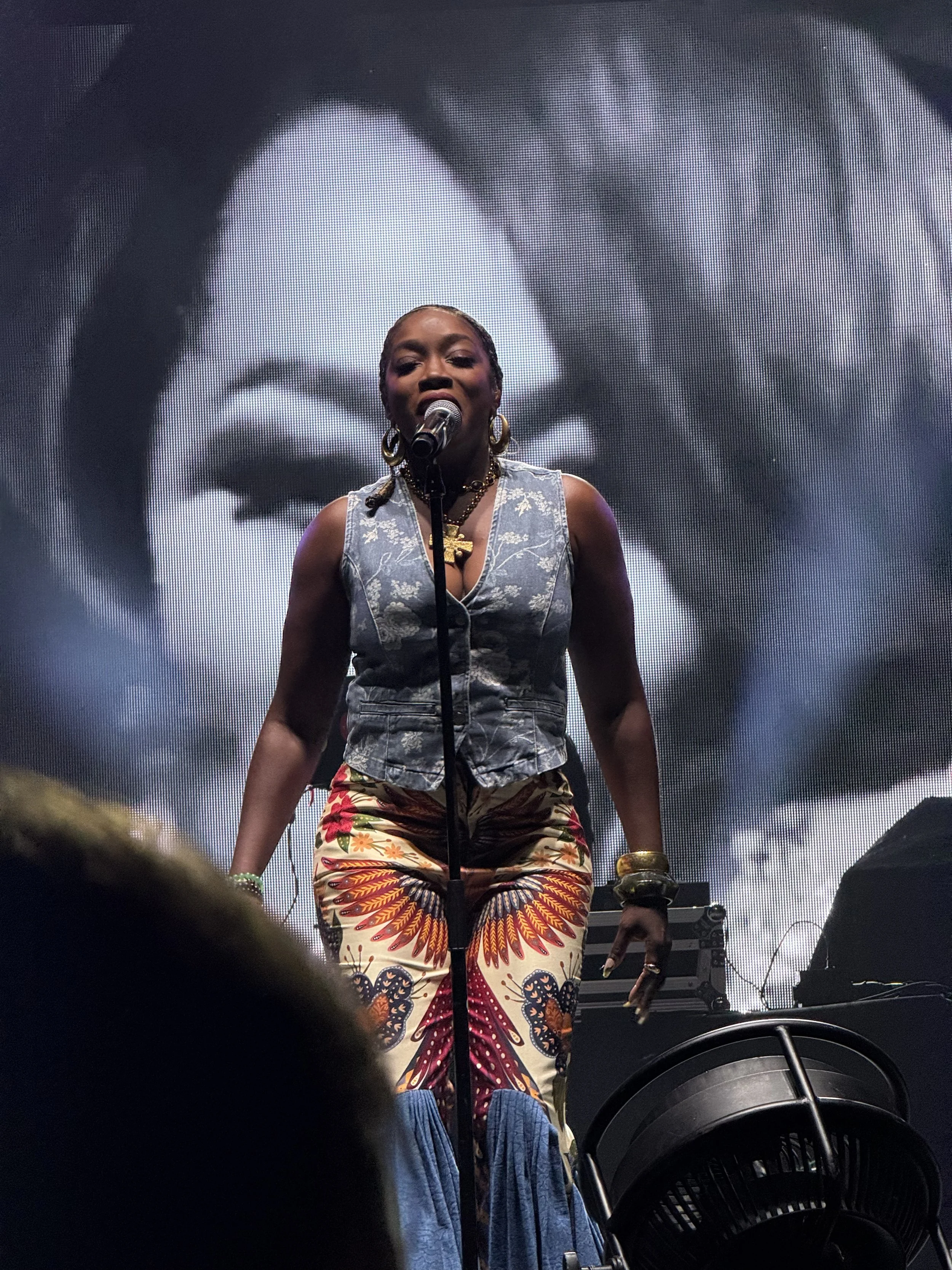 A woman performing on stage, standing in front of a large black-and-white portrait of a smiling person. The woman is wearing a patterned vest, colorful patterned pants, and gold jewelry, with a microphone in front of her.