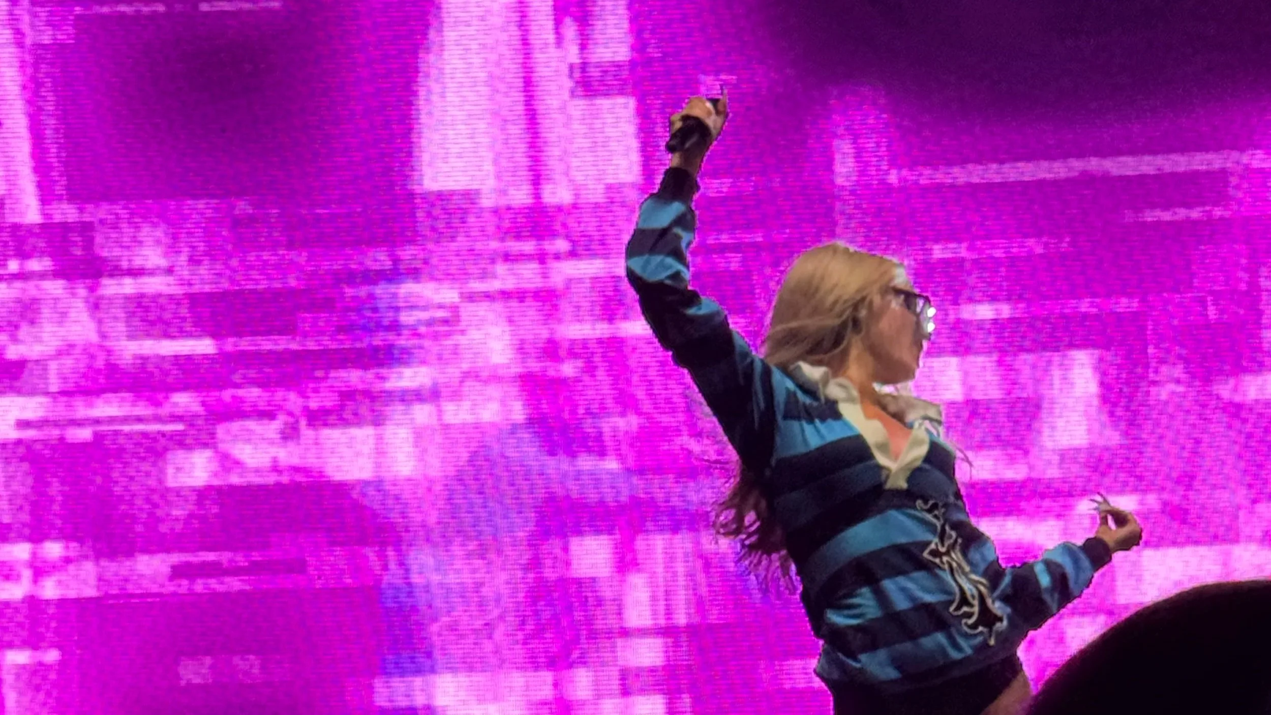 A woman with long hair and glasses, wearing a blue and gray striped shirt, is on stage holding up a microphone with her right hand. The background is a large pink and purple digital screen.