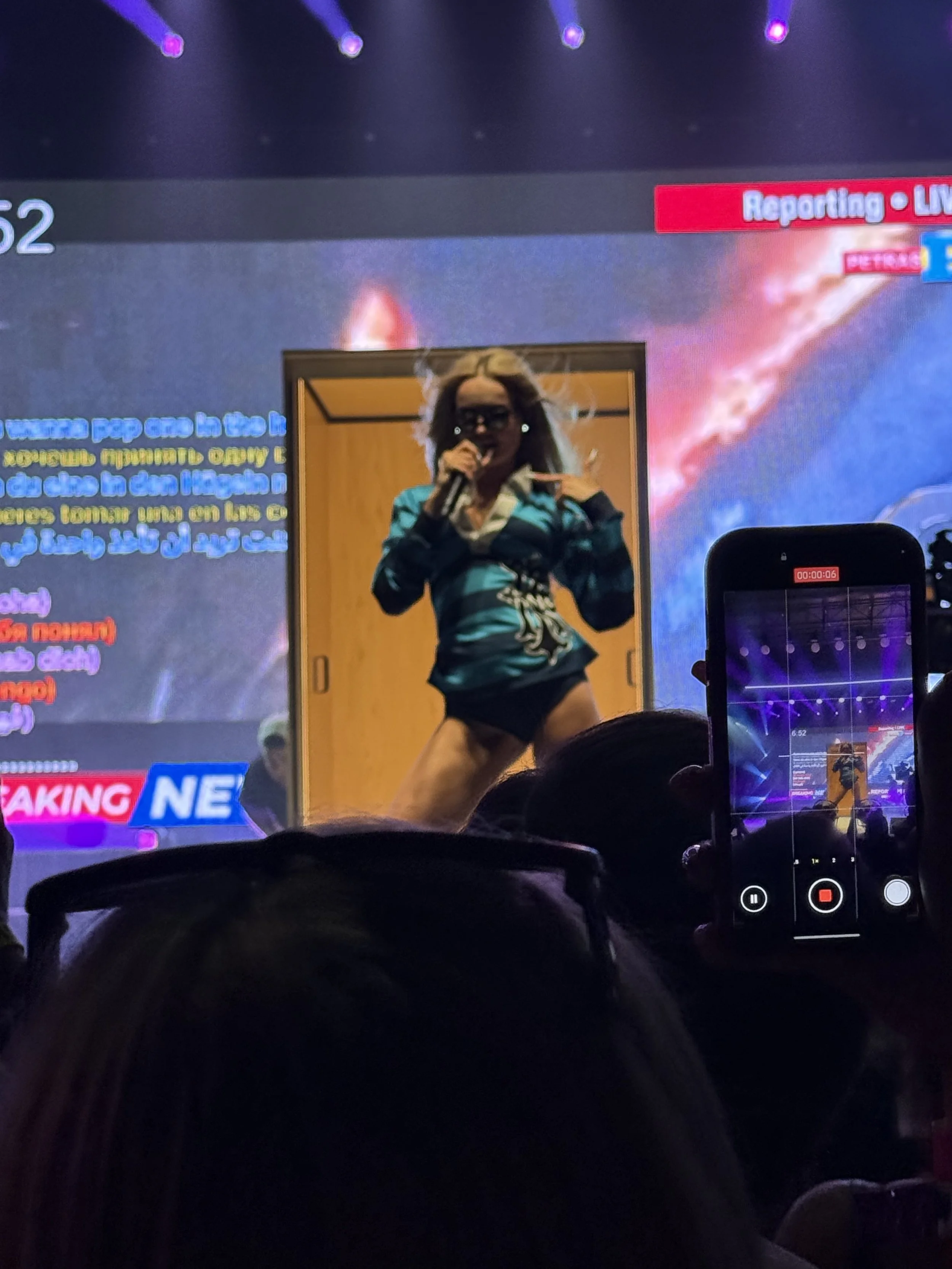 A woman in a green jacket and black shorts is dancing on stage, holding a microphone, in front of a large screen with colorful graphics and text, in a crowded event space with audience members taking photos.