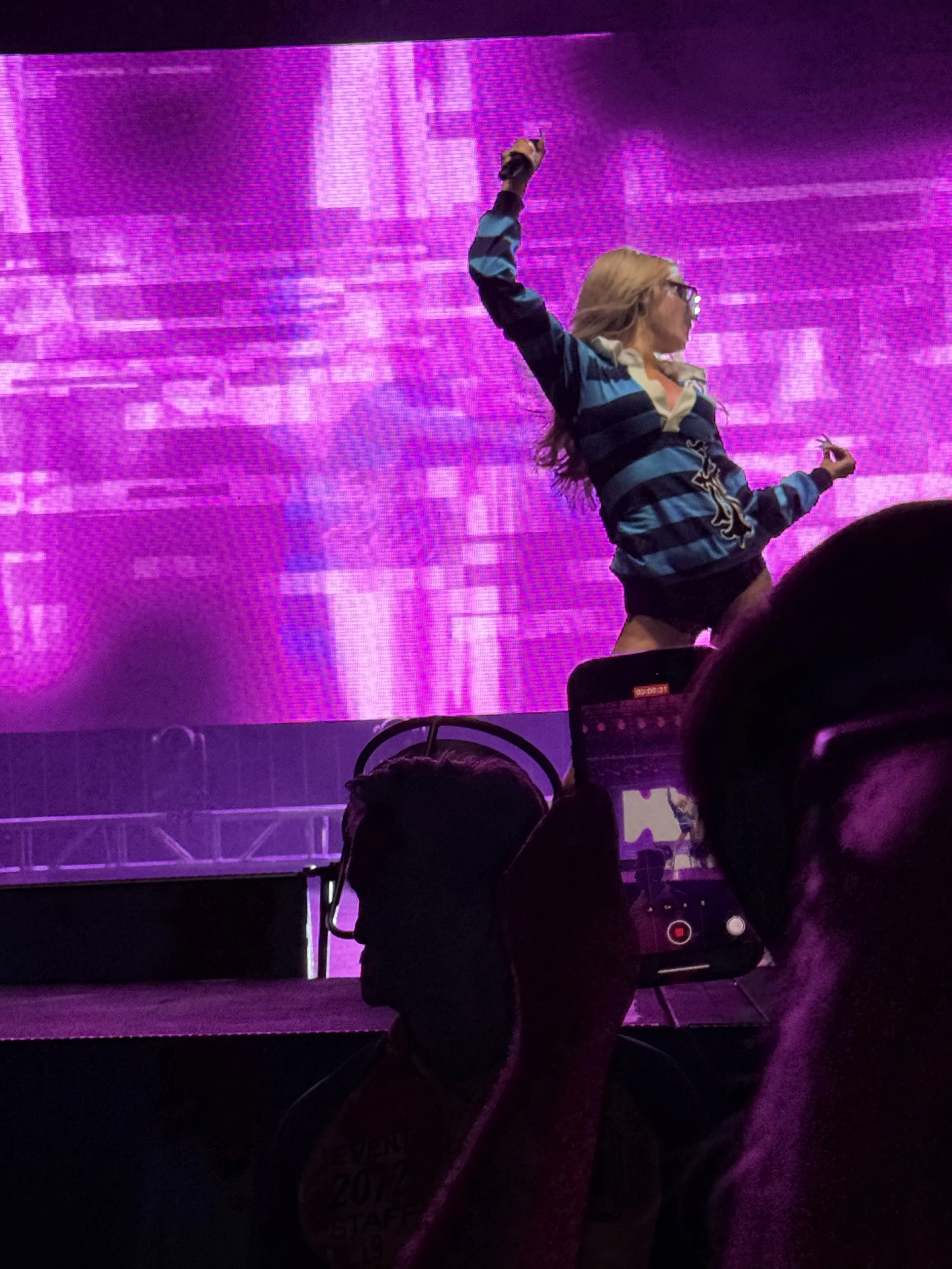 Person with long hair and glasses wearing a striped hoodie dancing or performing on a stage with a large bright purple digital screen in the background. Audience members are visible in the foreground.