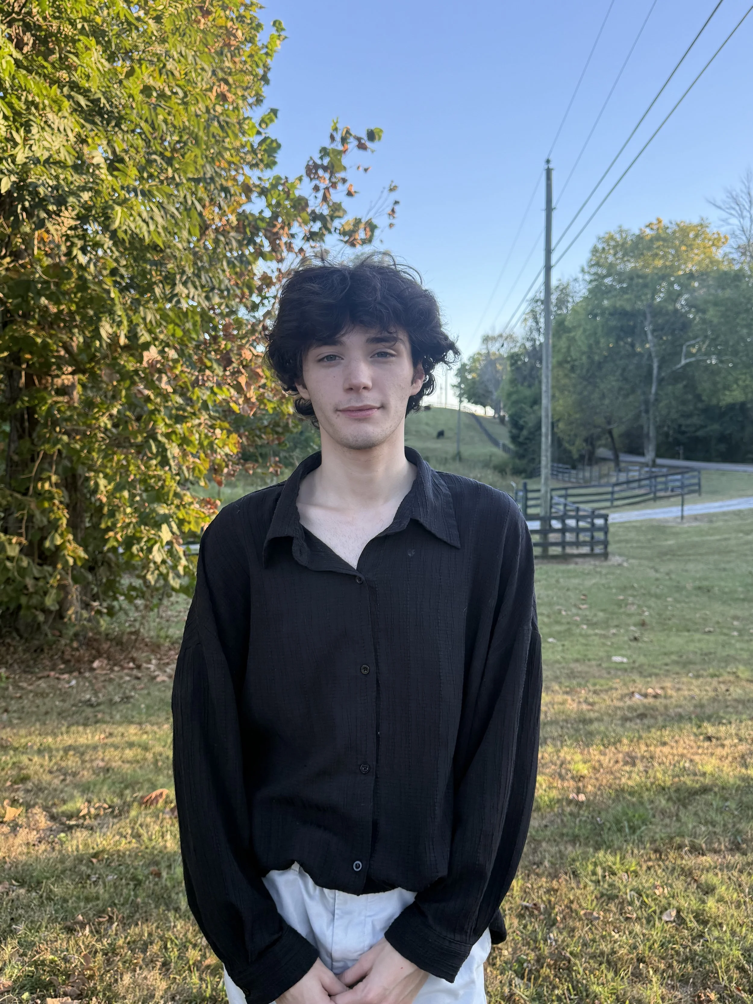 A young man with curly dark hair and fair skin standing outdoors on a grassy area during daytime. He is wearing a black button-up shirt over a white shirt, with his hands clasped in front of him. Behind him are trees with green and orange leaves, a wooden fence, and power lines under a clear blue sky.