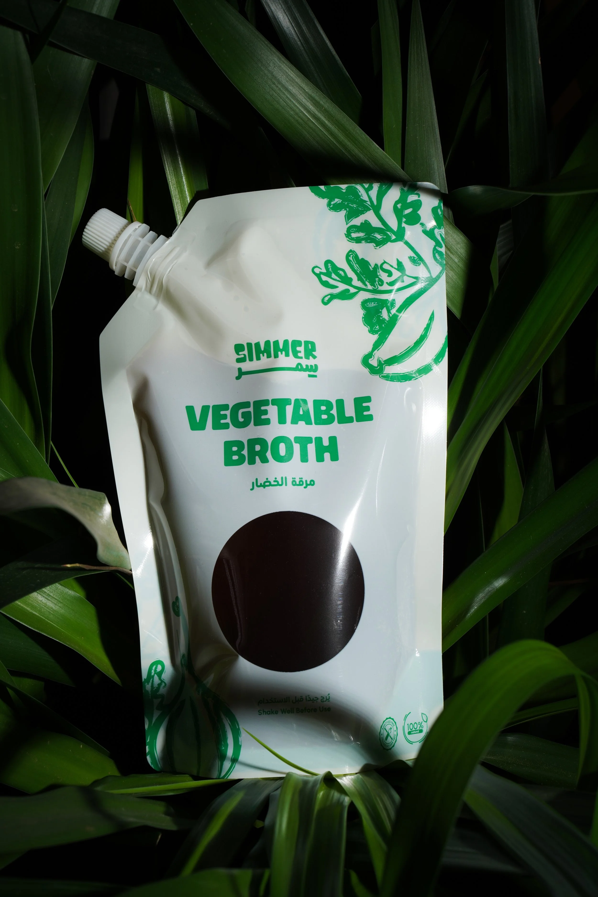 A pouch of vegetable broth amid green plant leaves, with some text in English and Arabic, and symbols indicating natural ingredients and recycling.