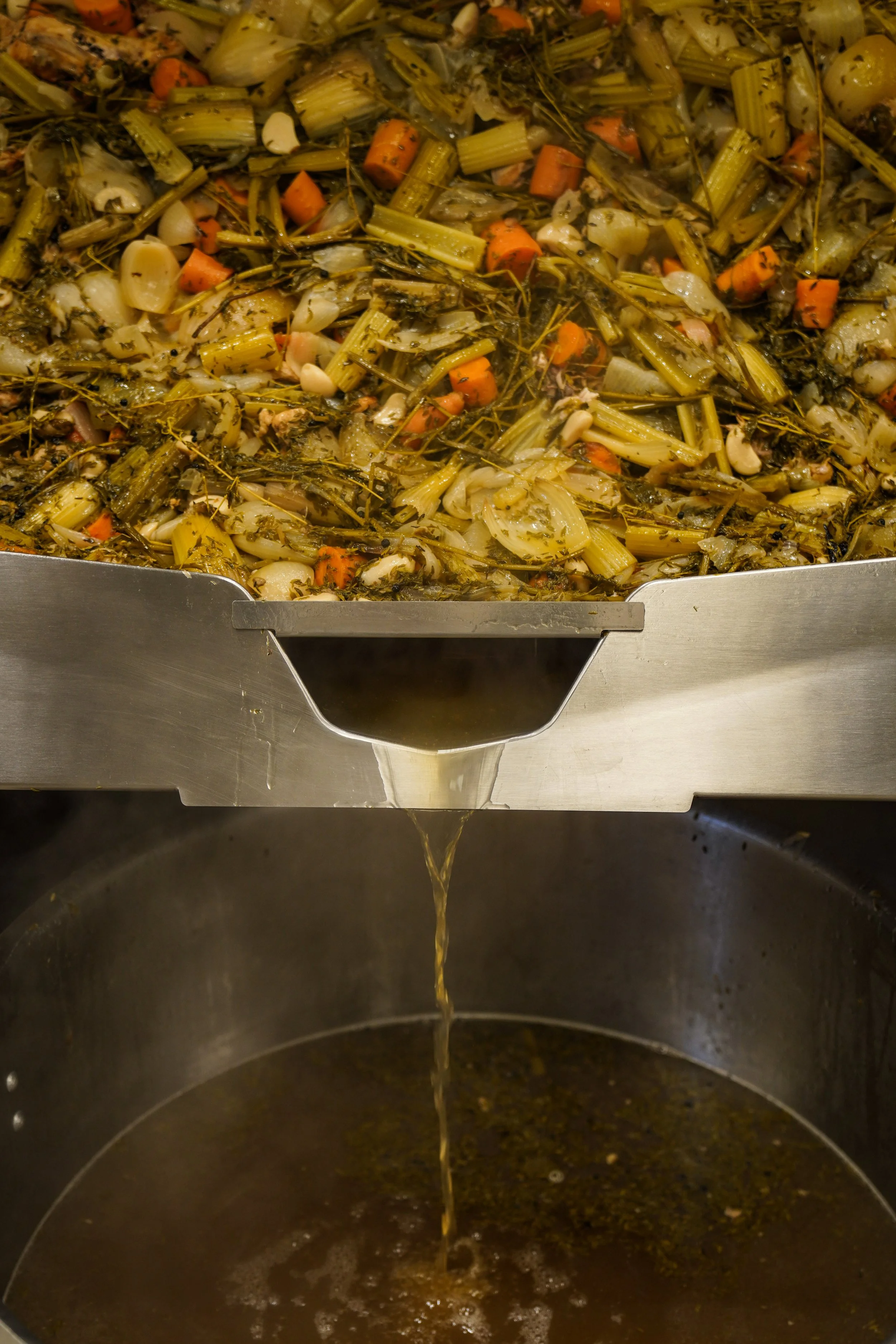 Vegetables like carrots, celery, onions, and garlic being cooked in broth or stock in a commercial kitchen or food processing setting.