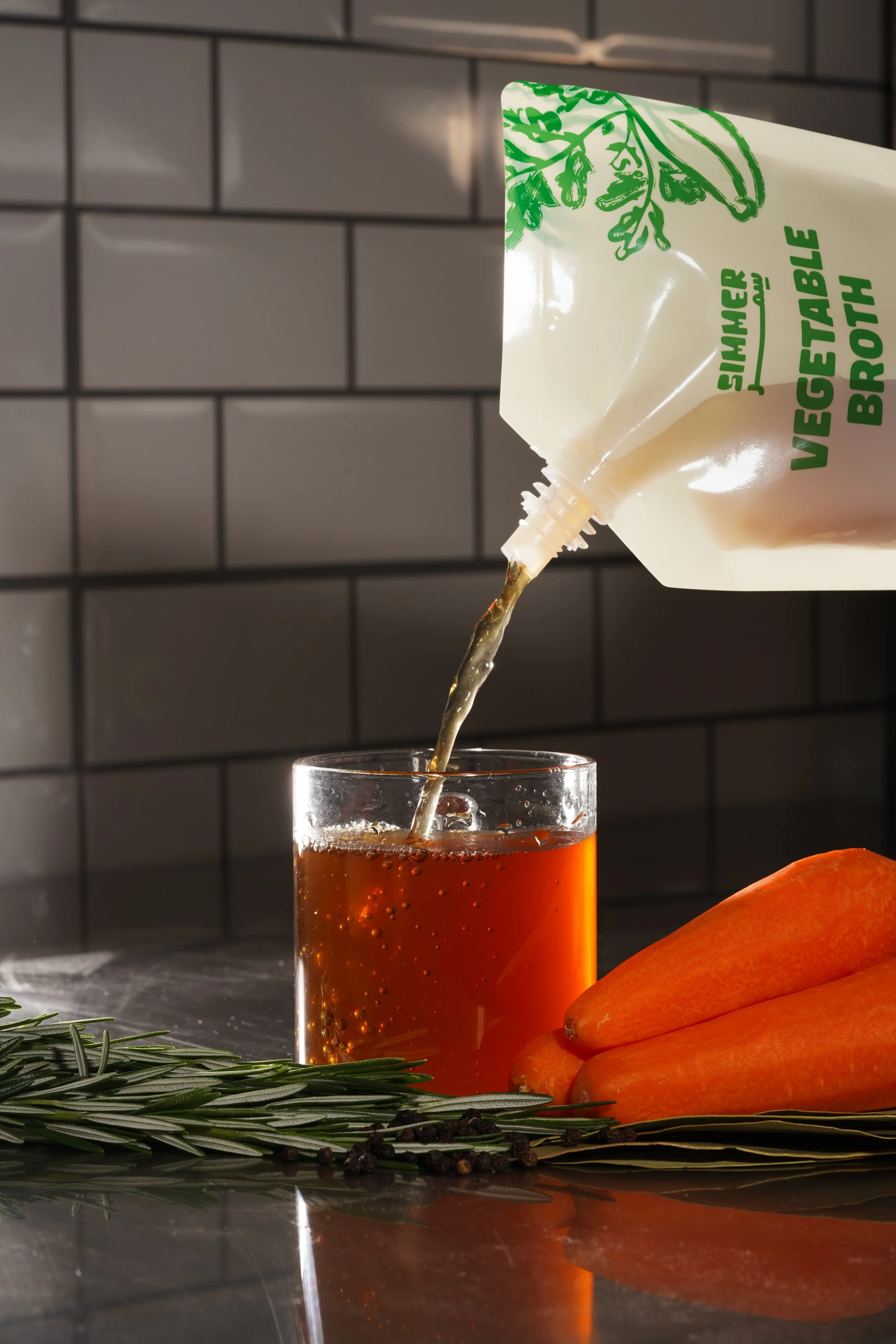 Pouring vegetable broth from a bag into a glass of dark liquid, with carrots, rosemary, and black peppercorns on a reflective surface.
