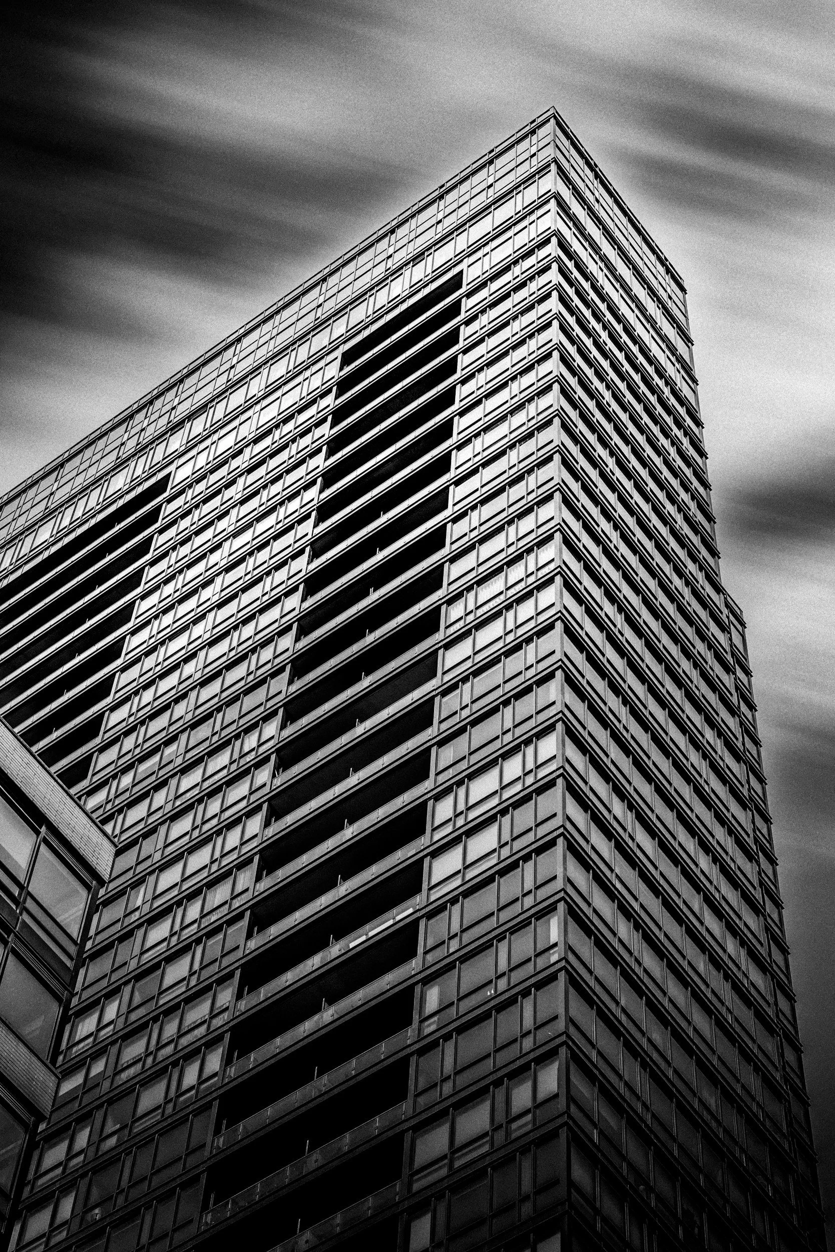 A tall, modern building with multiple glass windows and balconies, shot from a low angle against a cloudy sky in black and white.