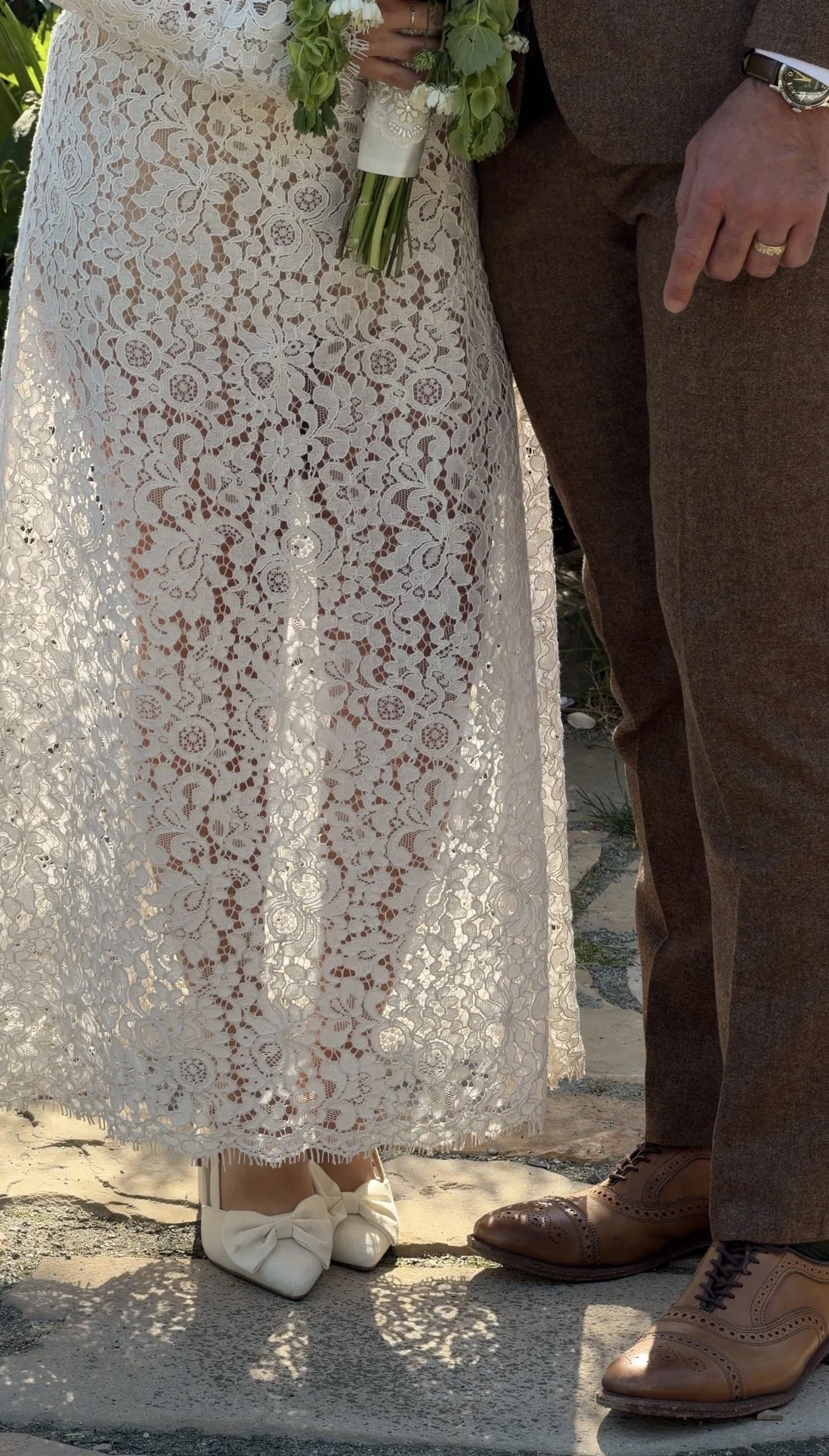 Close-up of a bride and groom standing close together, showing their legs and shoes. The bride wears white lace footwear with bows, and the groom wears brown leather brogue shoes. The bride holds a small bouquet of flowers.