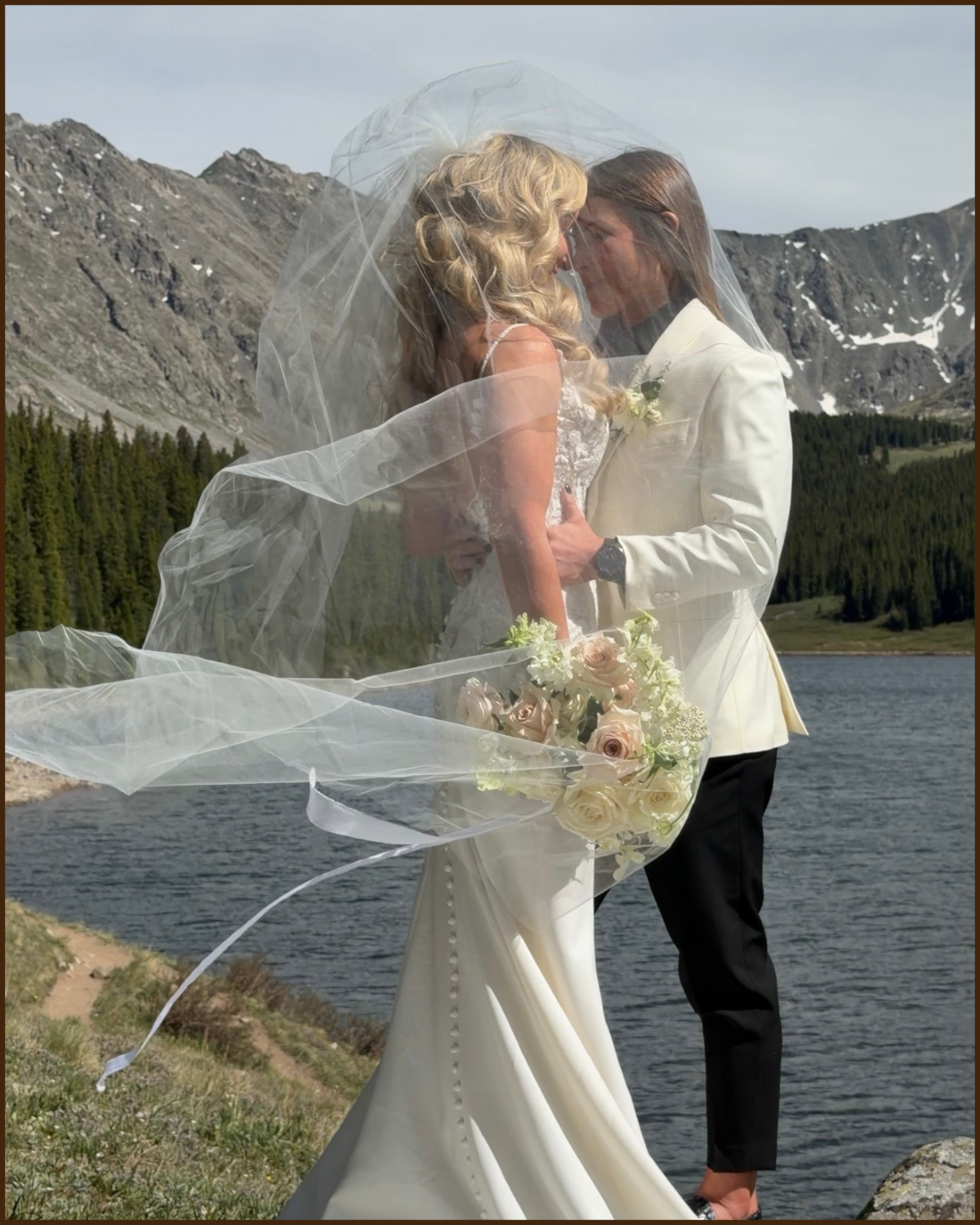 A bride and groom stand close together on a lakeshore, with mountains and a forest in the background, sharing a romantic moment. The bride is wearing a white wedding gown with a long train and veil, holding a bouquet of white and blush roses, and the groom is dressed in a white tuxedo jacket and black pants.