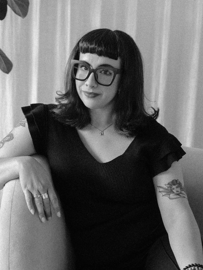 A woman with dark hair and bangs, wearing large glasses, a black top with ruffled sleeves, a necklace, and tattoos on her arms, sitting on a couch.