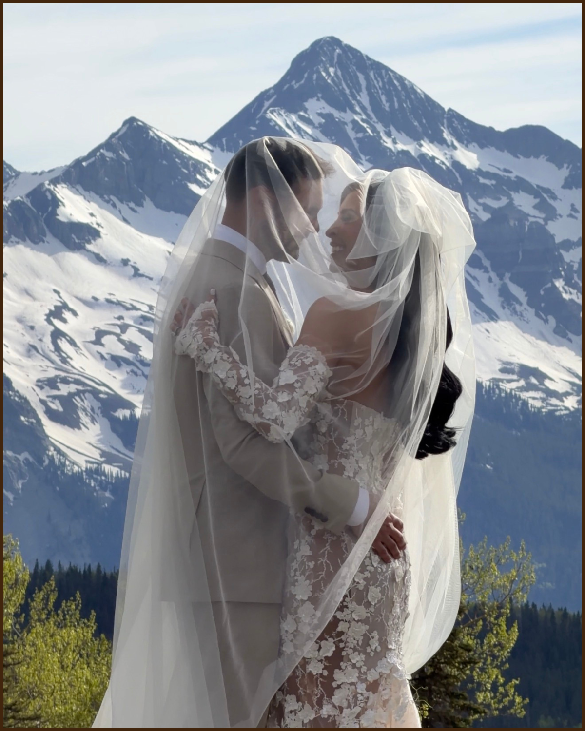 A bride and groom embracing in wedding attire with snow-capped mountains in the background.