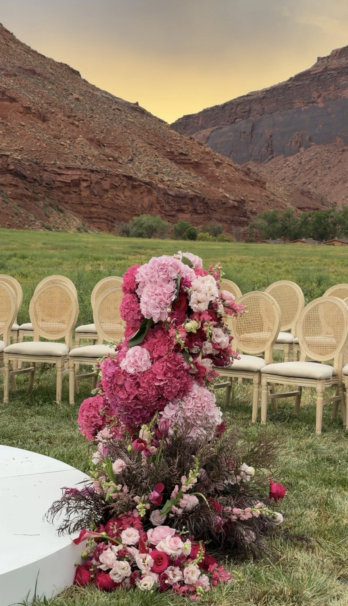 A floral arrangement consisting of pink, red, and white flowers in front of empty chairs outdoors with mountain cliffs in the background at sunset.