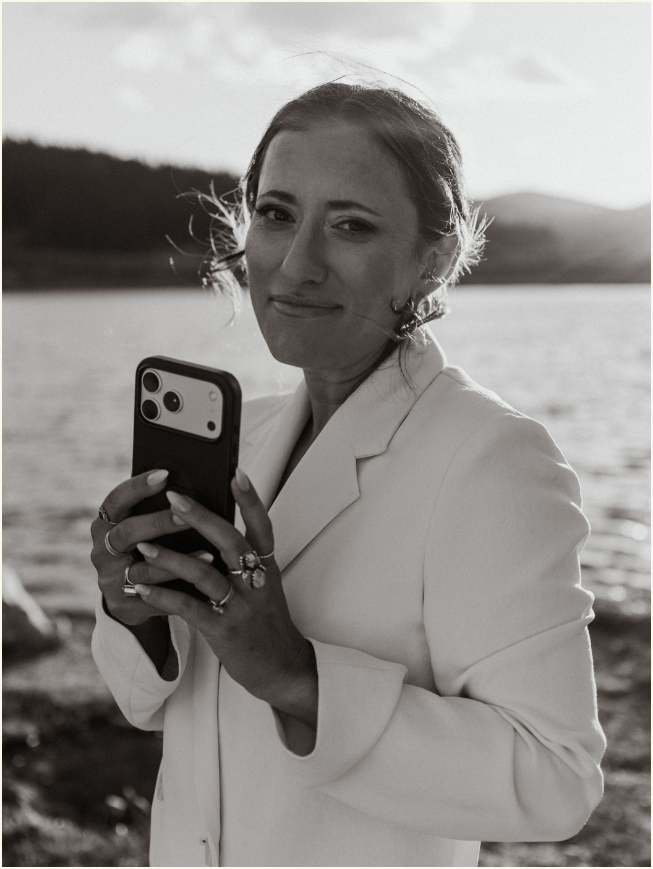 A woman in a white blazer holding a smartphone near a body of water, with a scenic outdoor background, smiling gently at the camera.