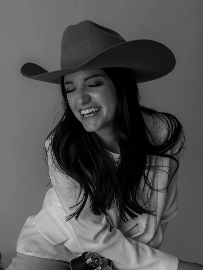 Black and white photo of a woman smiling with her eyes closed, wearing a wide-brimmed hat and an off-the-shoulder top.