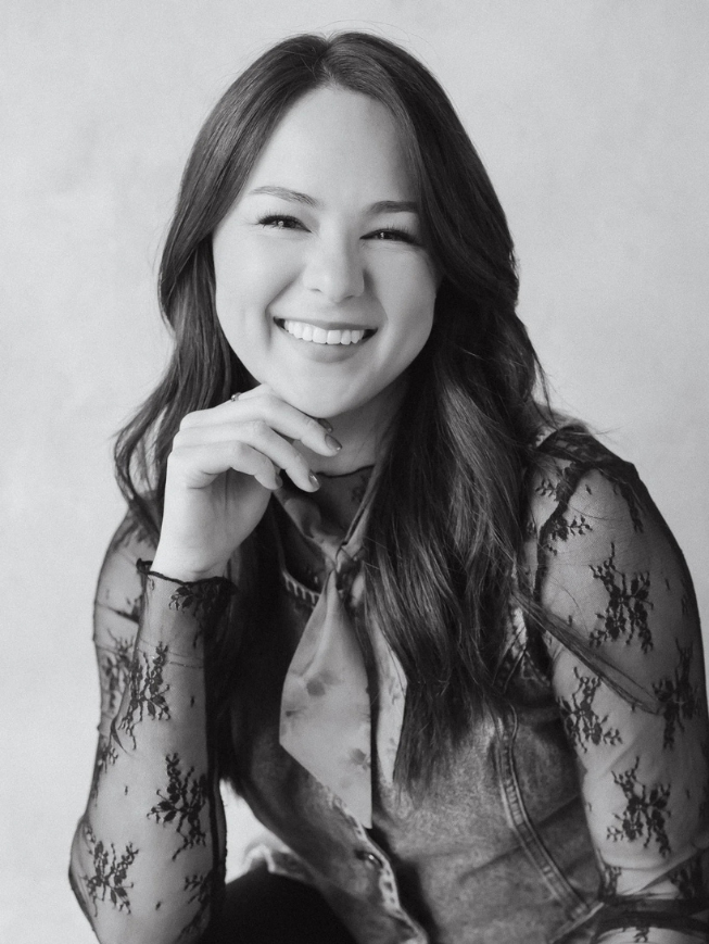 Black and white portrait of a smiling young woman with long hair, wearing a lace patterned blouse and denim jacket, posing with her hand near her chin.