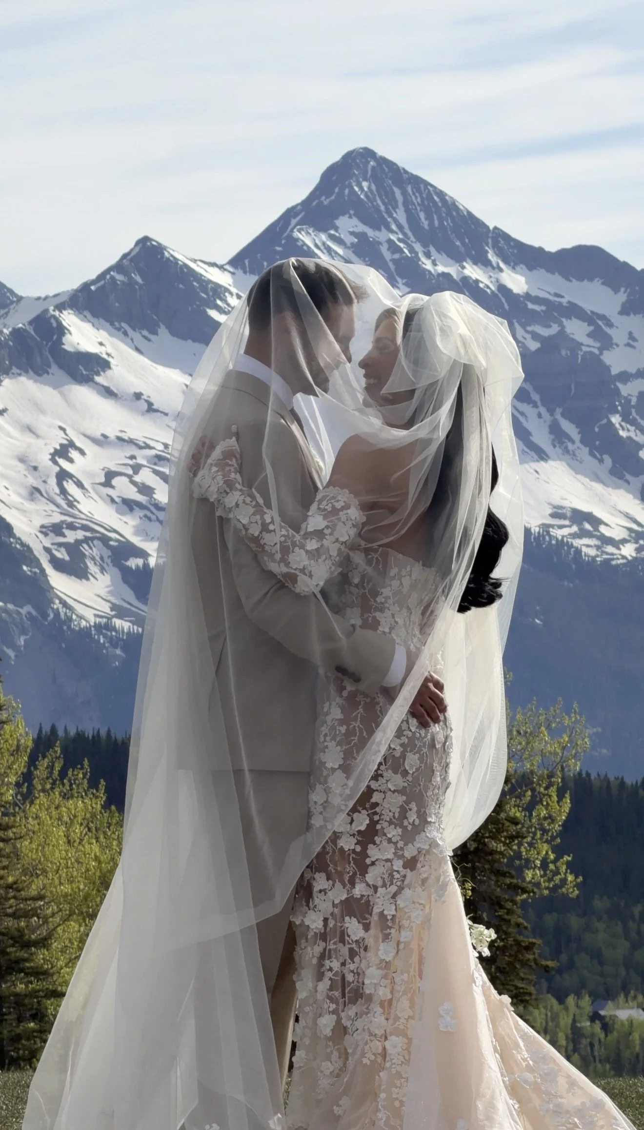 A bride and groom standing close together beneath a veil, with snow-capped mountains in the background, during a wedding photoshoot.