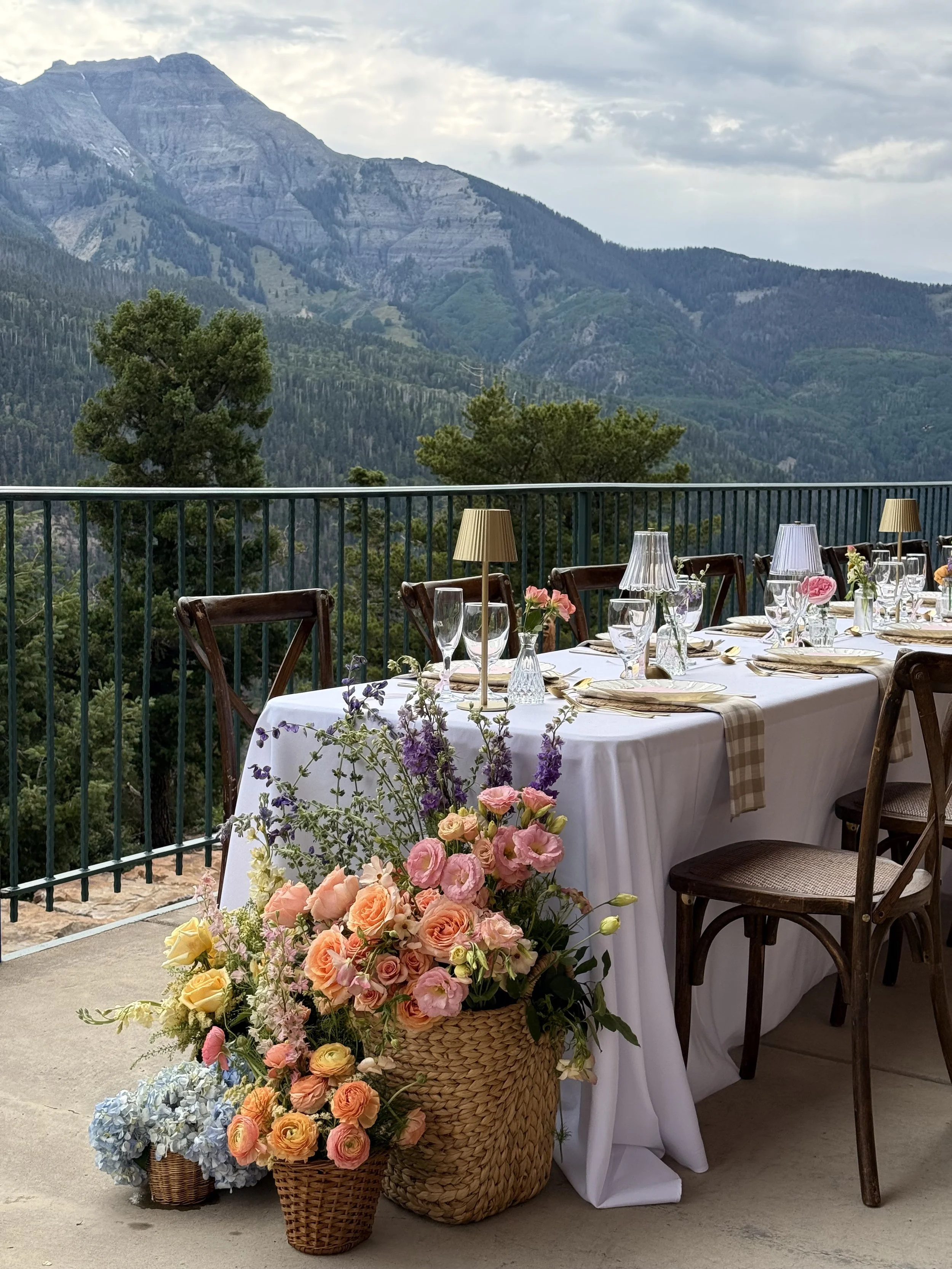 A decorated outdoor dining table with floral centerpieces, glassware, and lamps, set against a mountain landscape with trees and clouds.