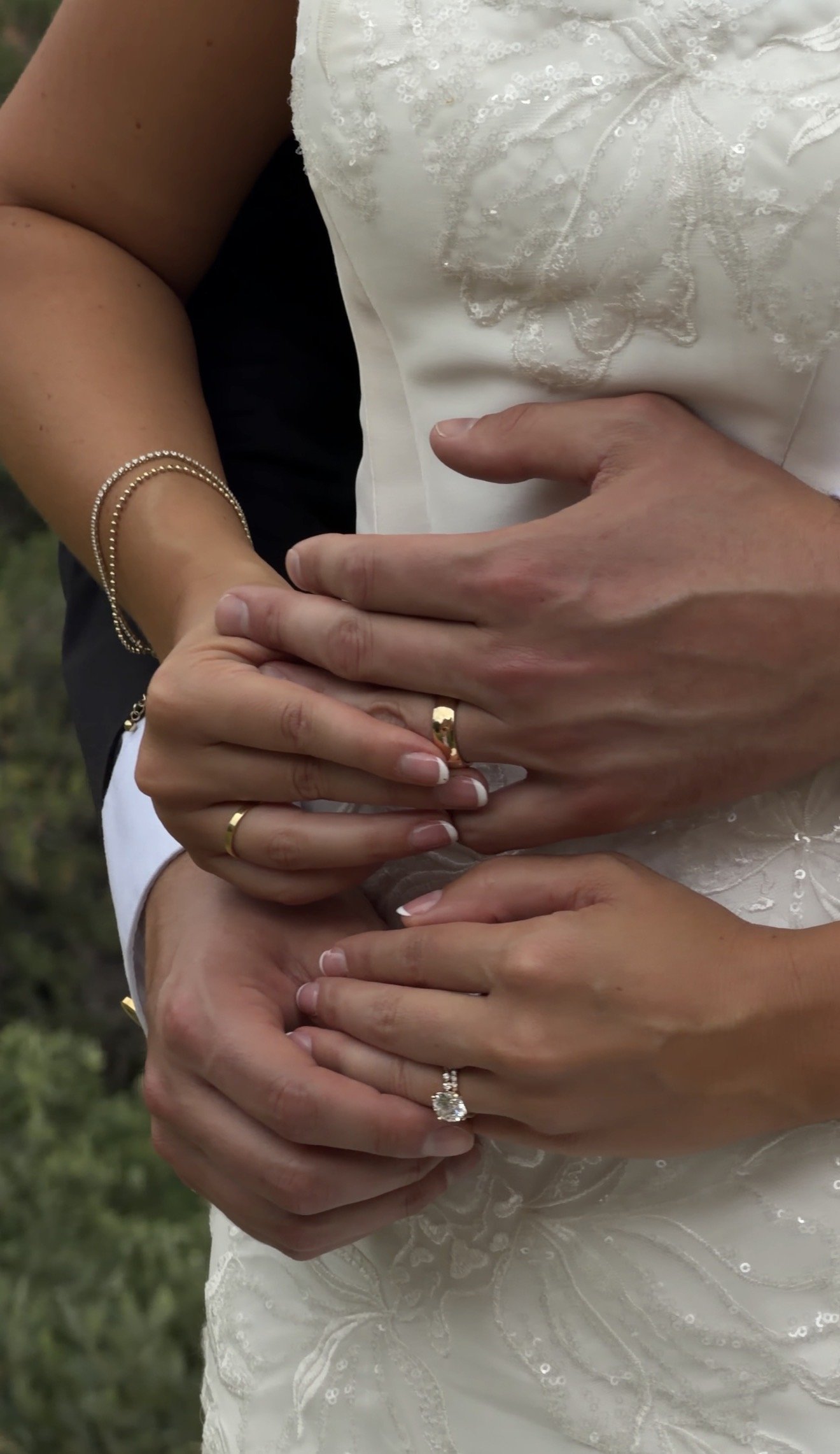 A close-up of a couple's hands intertwined, with the woman wearing a wedding dress and engagement ring, and the man wearing a wedding band.
