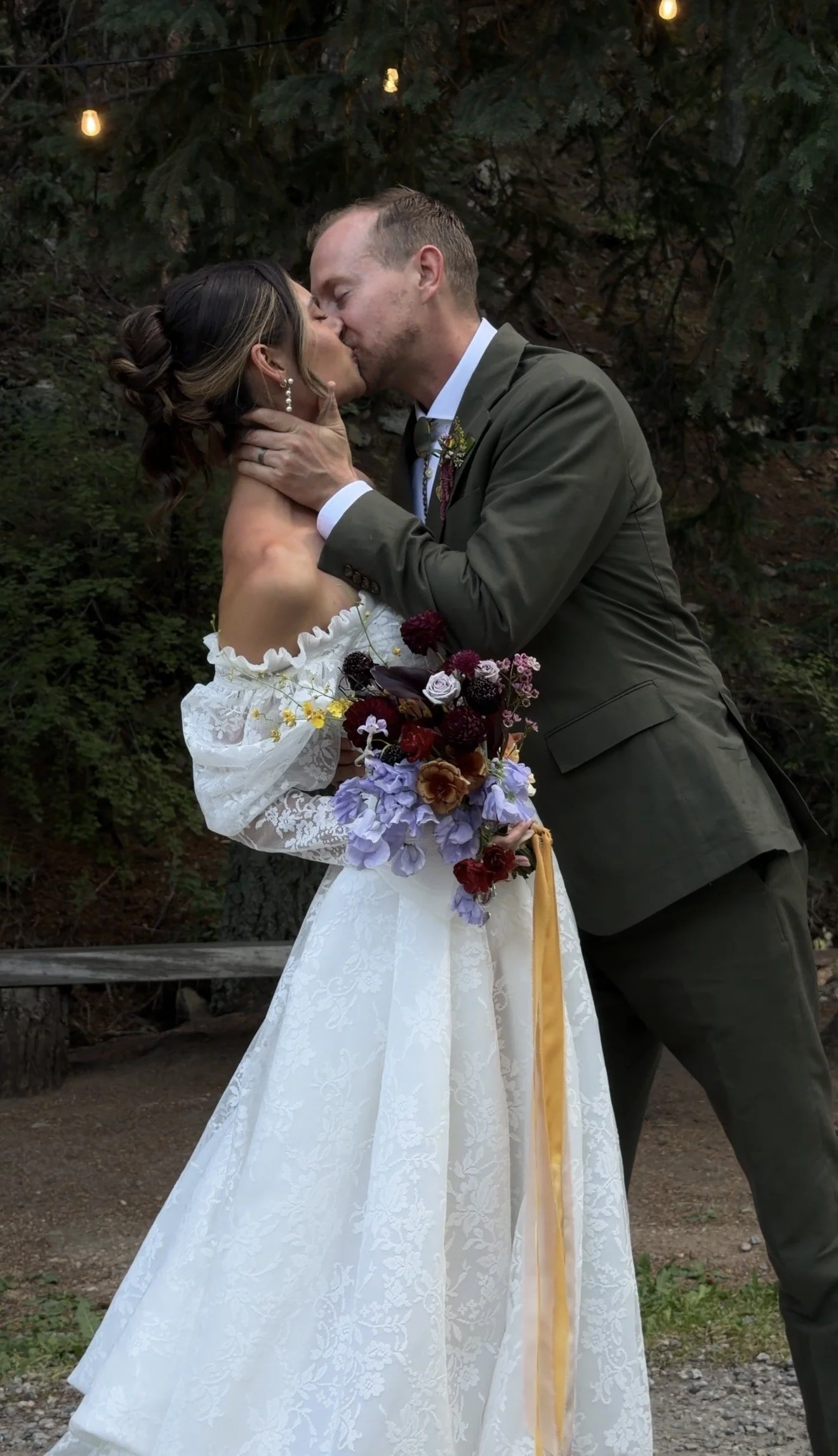 A couple in wedding attire kissing outdoors, with a woman holding a bouquet of purple, red, and yellow flowers. The man is wearing a dark green suit and the woman is wearing a white lace off-shoulder dress, with trees and hanging lights in the background.
