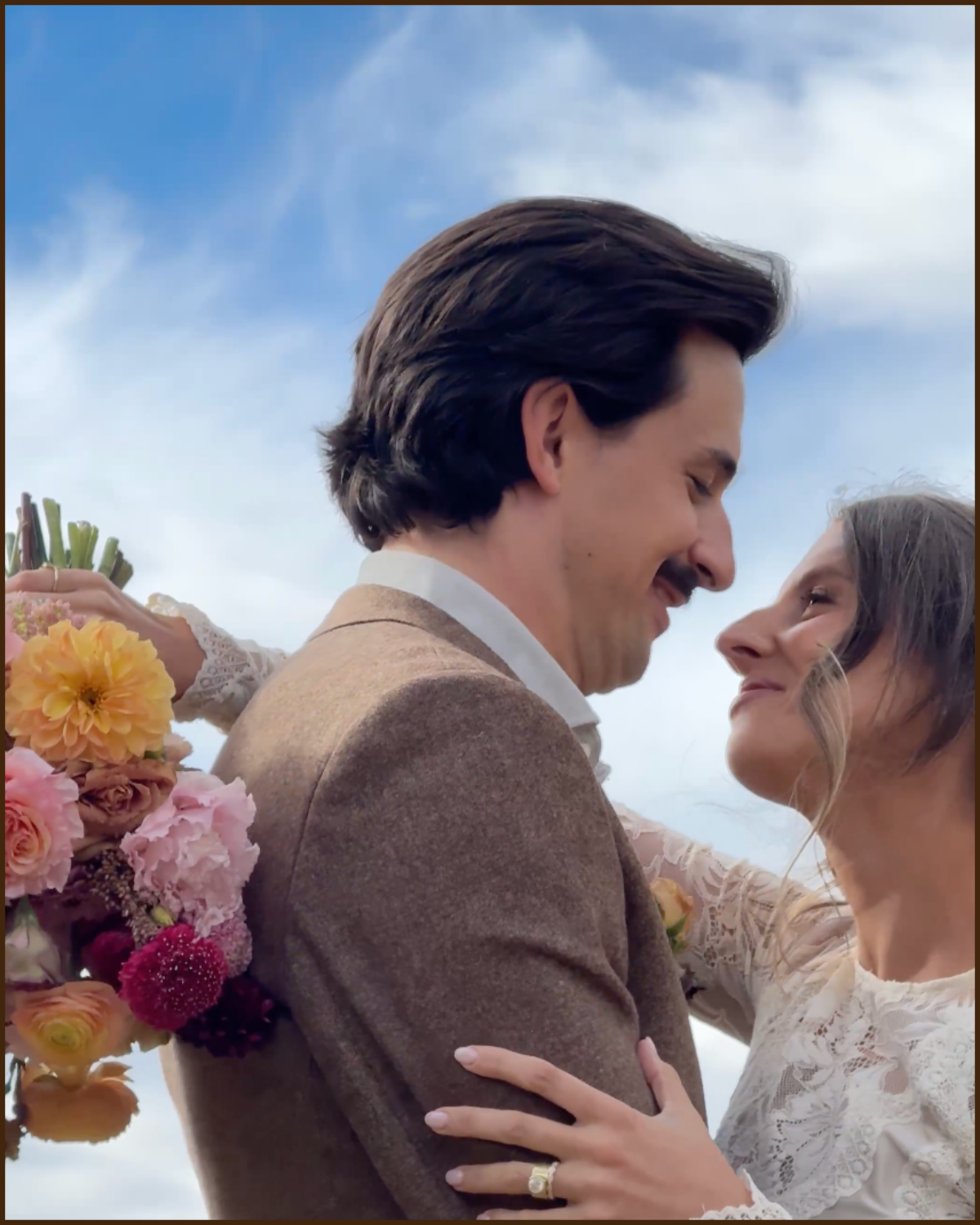 A man and woman are embracing and smiling at each other outdoors against a blue sky, with the woman holding a bouquet of flowers.