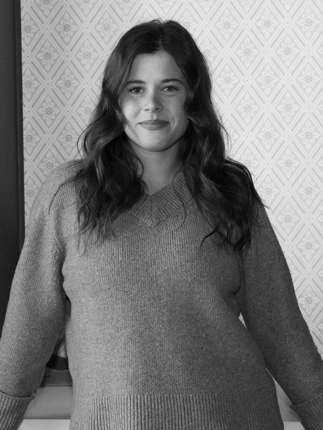 Black and white photo of a young woman with long wavy hair wearing a sweater, smiling slightly, standing indoors in front of a patterned wallpaper.