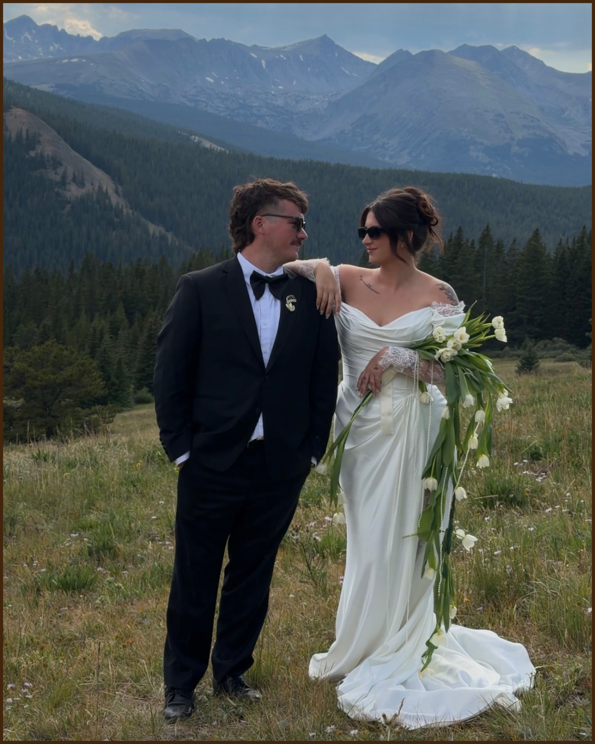 A couple in wedding attire standing outdoors in a grassy field with mountains and a forest in the background. The groom is wearing a black tuxedo with a bow tie, and the bride is in a white wedding dress holding a large bouquet of white flowers.