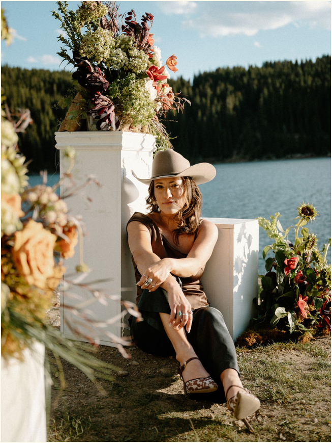 A woman sitting outdoors near a body of water, surrounded by colorful flowers and greenery, wearing a wide-brimmed hat and casual clothing.