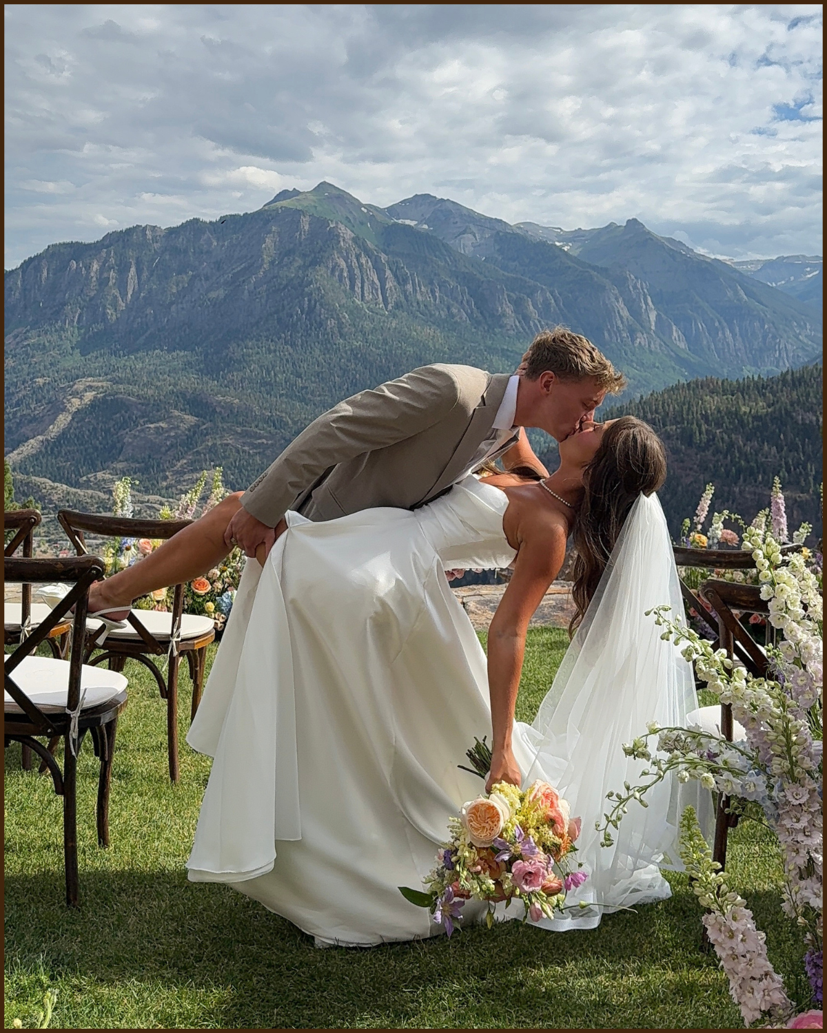 A newlywed couple sharing a kiss during their outdoor wedding ceremony with mountainous scenery in the background, surrounded by flowers.