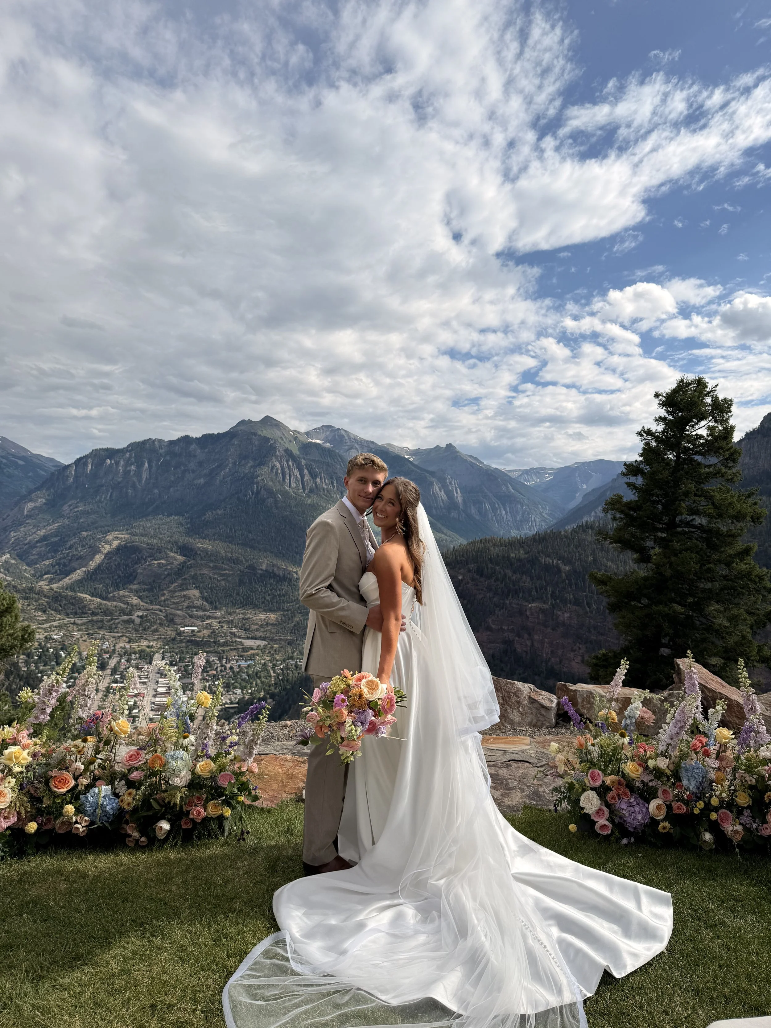 A bride and groom in wedding attire standing outdoors with mountains, trees, and a sky with clouds in the background, surrounded by colorful flowers.