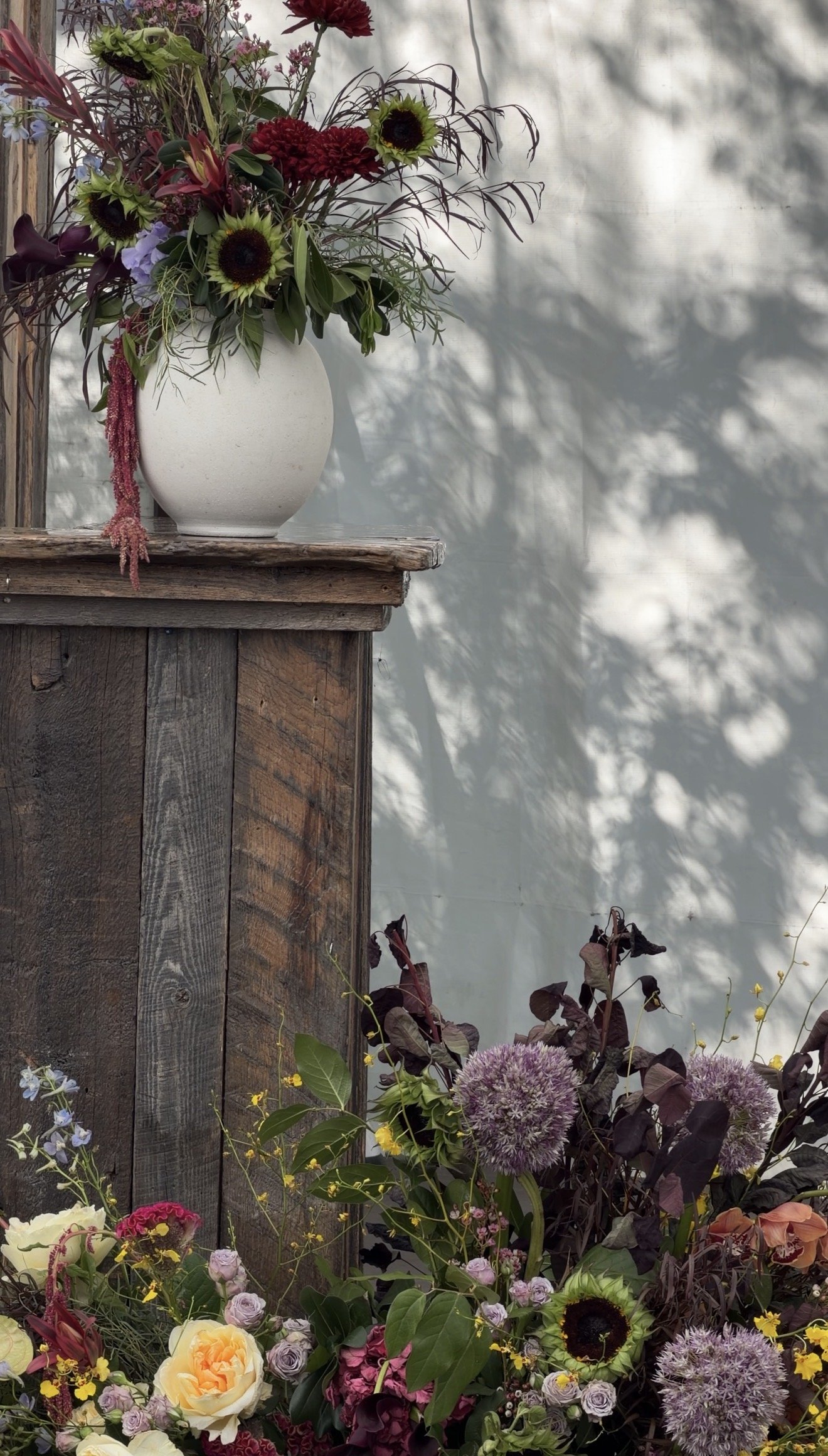A white ceramic vase with a bouquet of colorful flowers, including sunflowers, red, purple, and pink flowers, on a rustic wooden surface with some shadows from nearby trees or plants.