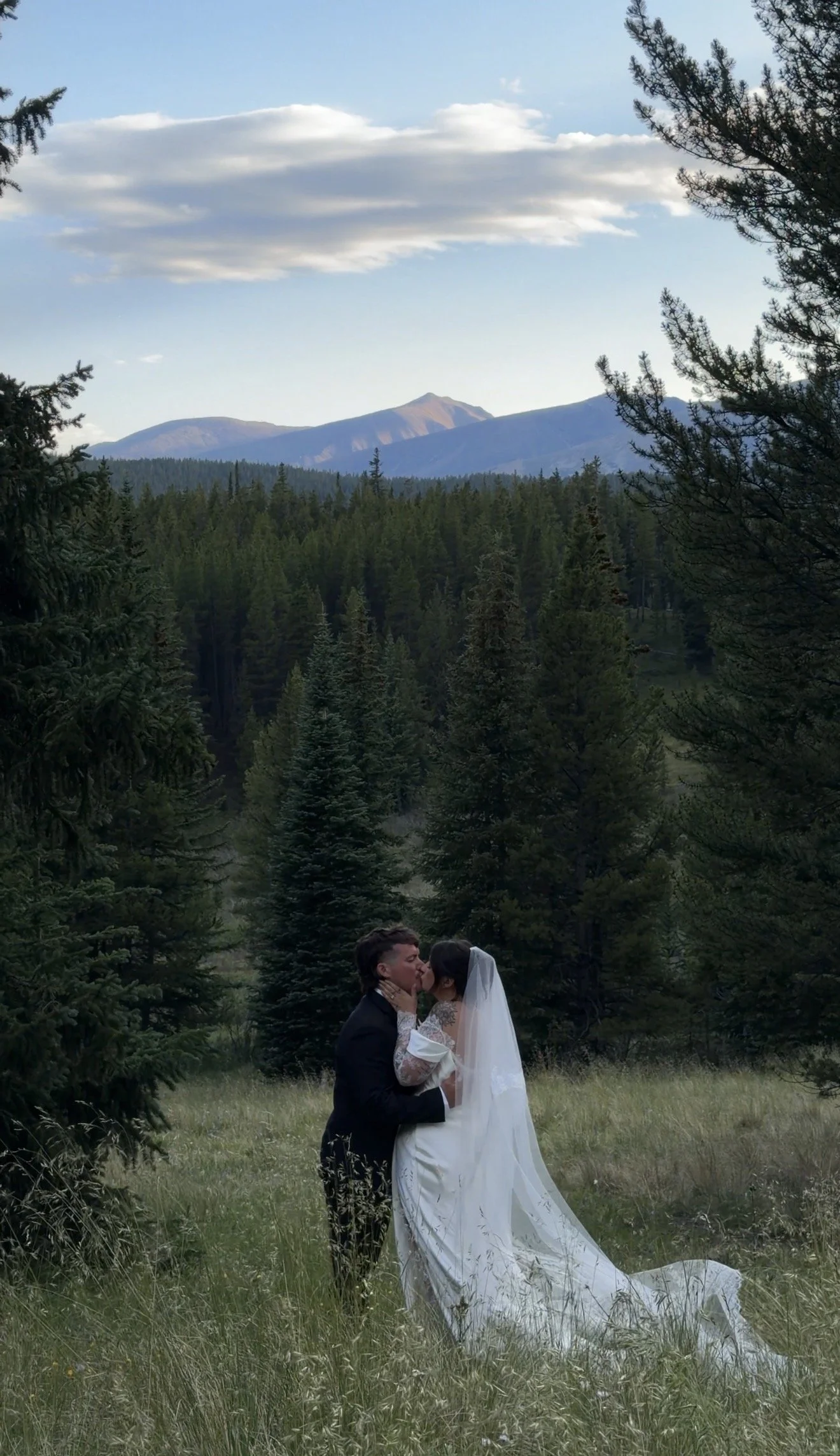 A bride and groom sharing a kiss in a forest clearing with mountains and a blue sky in the background.