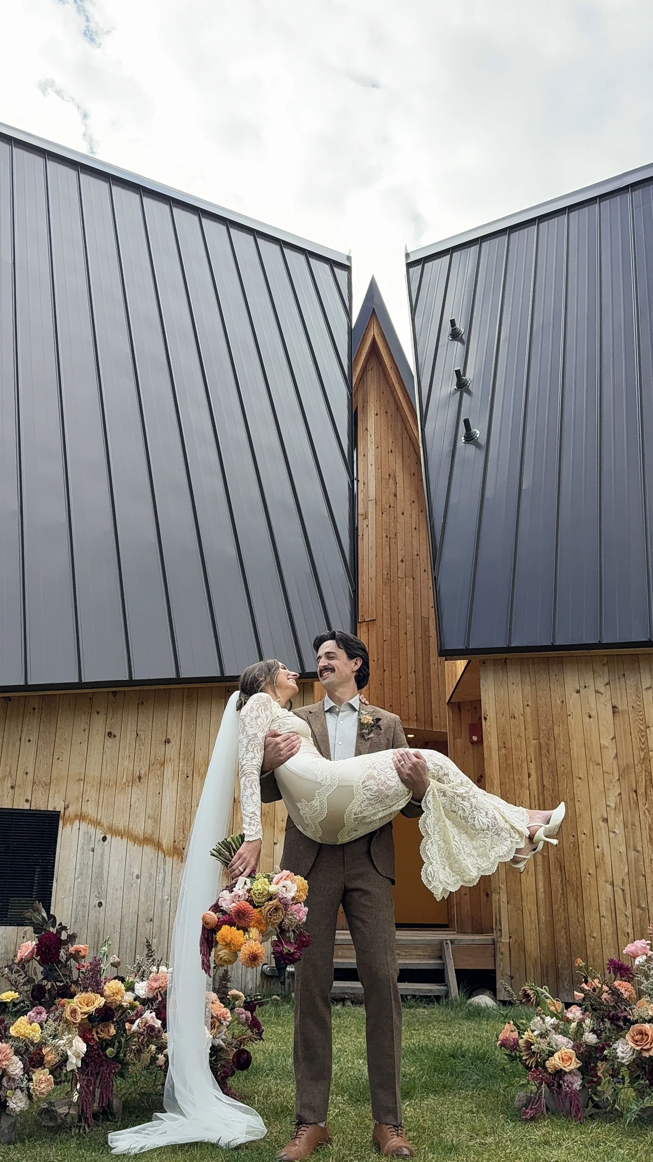 A groom carrying a bride in front of a modern wooden and metal building, surrounded by colorful flowers, during a wedding celebration.