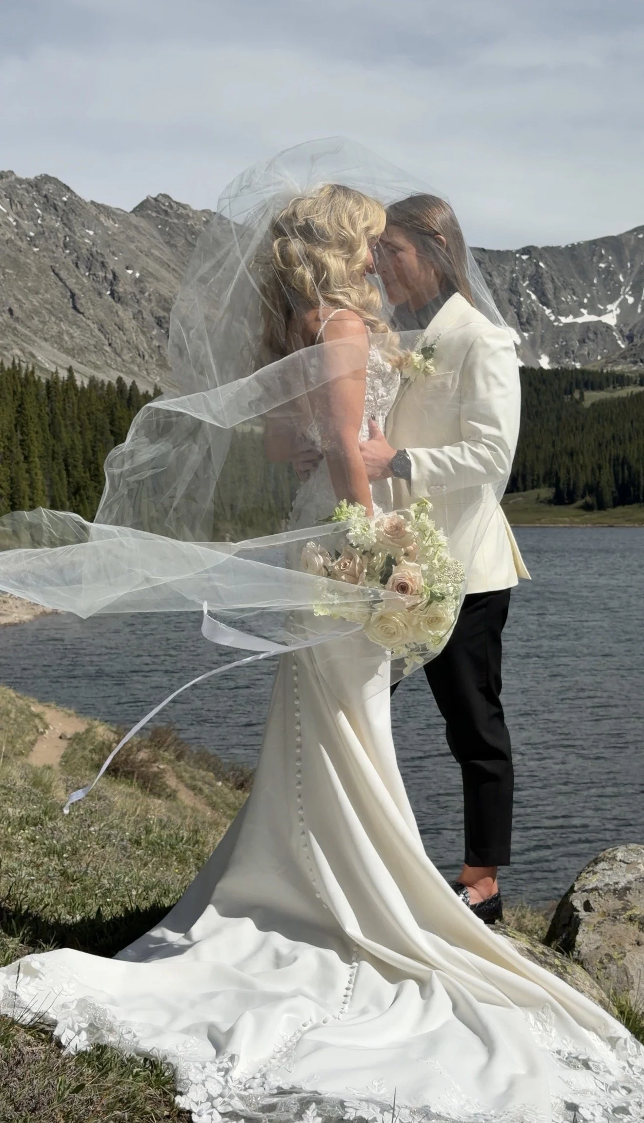 A bride and groom stand closely together outdoors by a lake with mountainous terrain in the background. The bride wears a white gown with a long train and has blonde, curly hair. The groom wears a white jacket, black pants, and loafers, with long hair. The bride holds a bouquet of white and blush roses. Their faces are close together beneath a sheer veil, sharing an intimate moment.