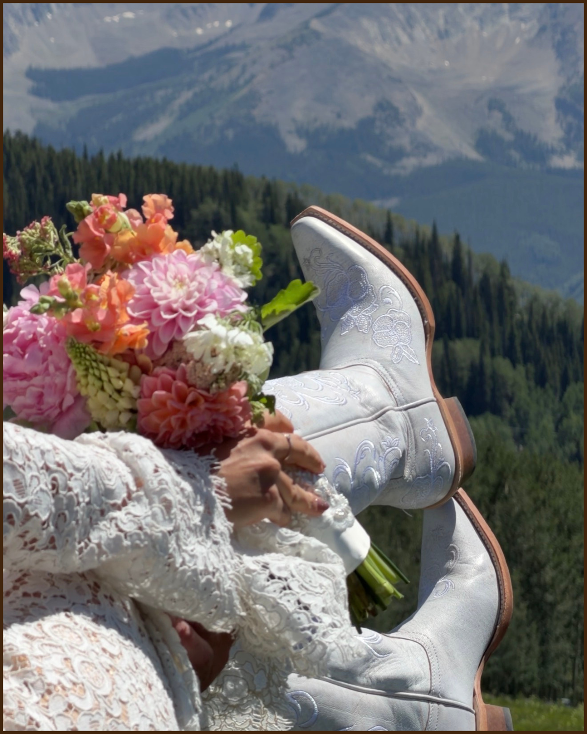 A person in white embroidered cowboy boots and a white lace dress holding a bouquet of pink and white flowers, with a scenic mountain landscape in the background.