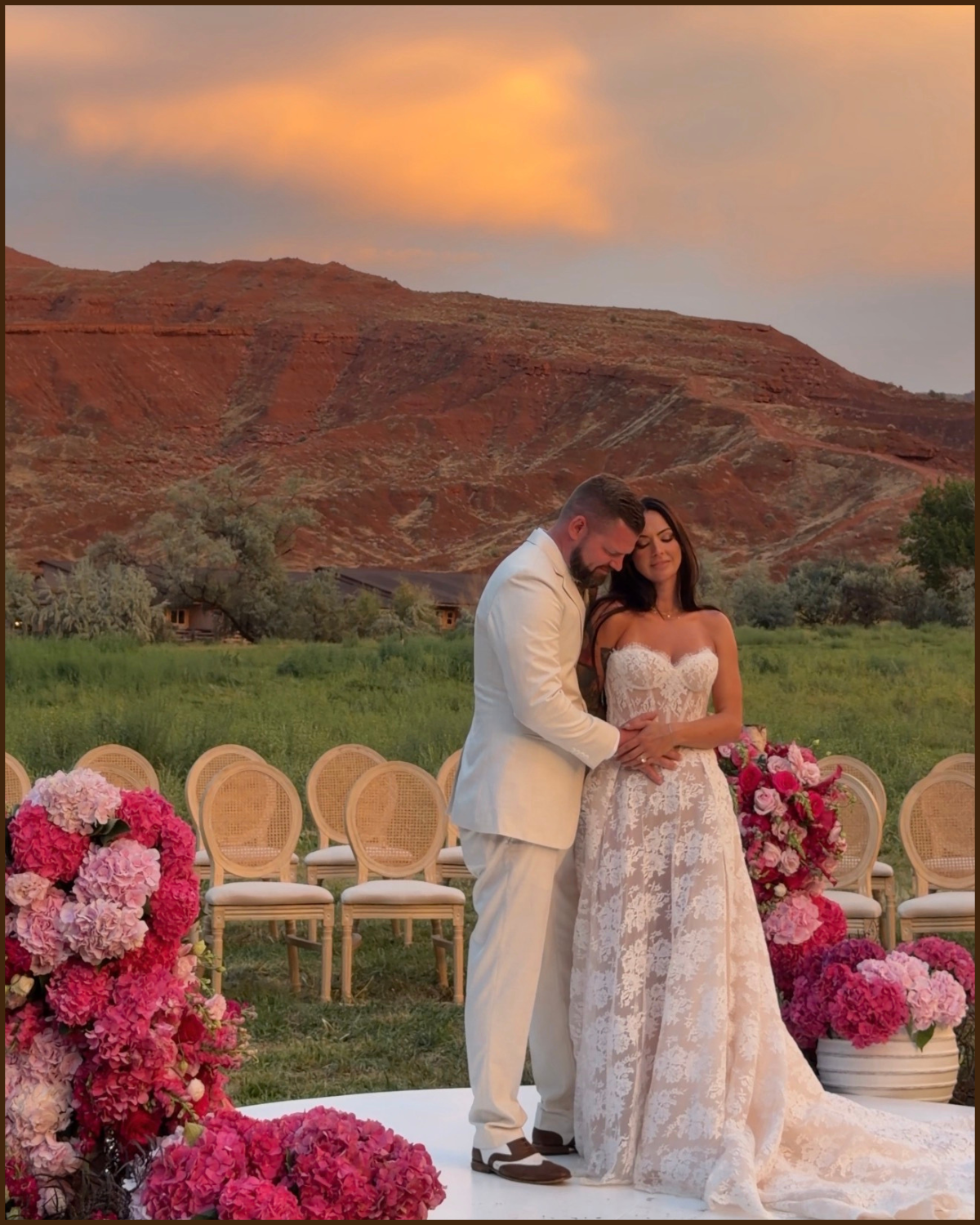 A bride and groom embrace during an outdoor wedding ceremony at sunset, with pink and purple flowers arranged around them and a scenic mountain landscape in the background.