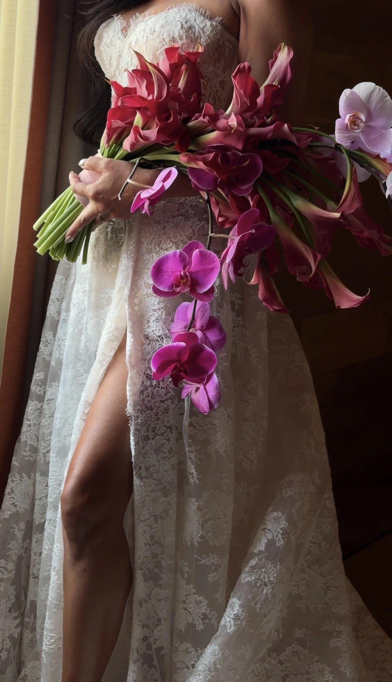 A woman wearing a white lace dress holding a bouquet of pink and red calla lilies and orchids.