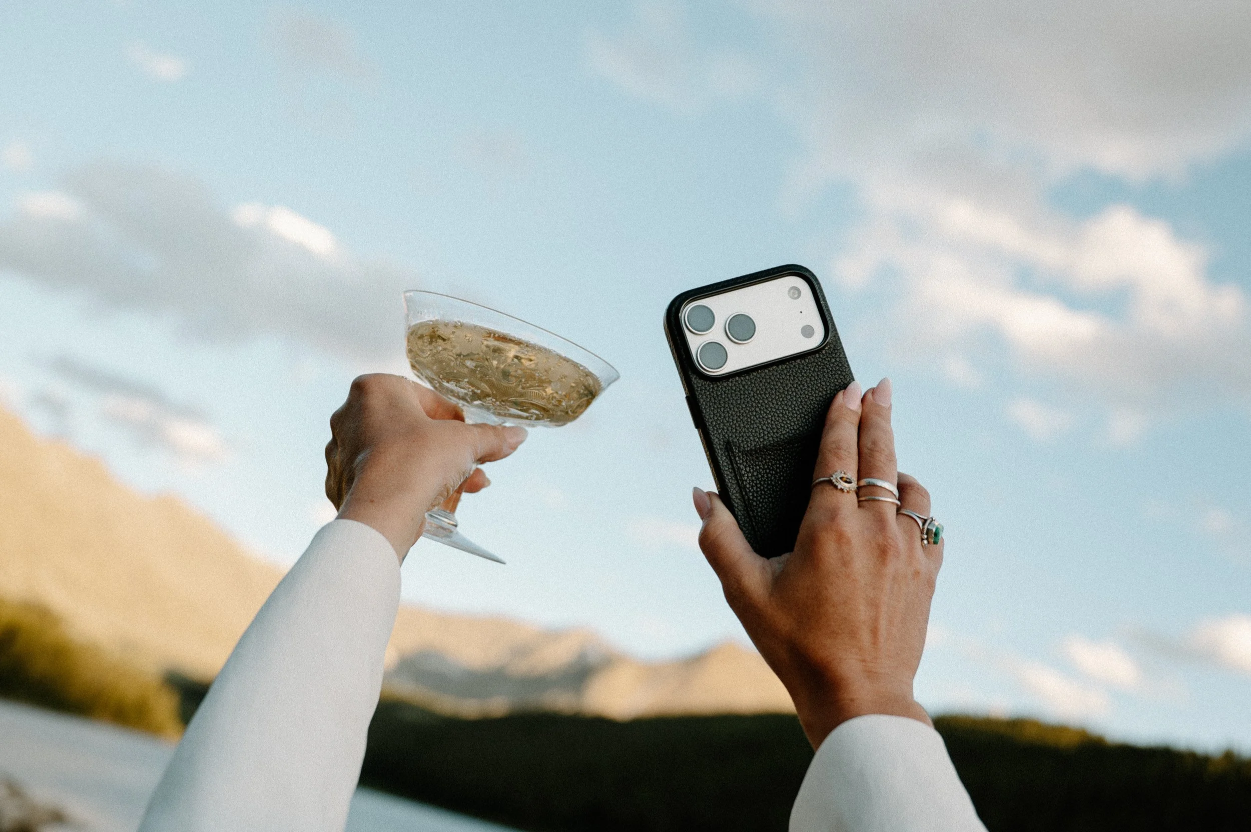 Person holding a champagne glass in one hand and a smartphone in the other, with two other people’s hands visible in the background, all against a sky with clouds.