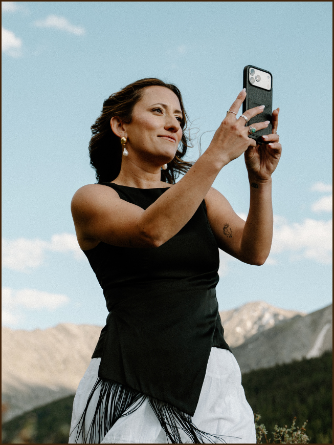Woman in black sleeveless top taking a selfie outdoors with mountains and blue sky in the background.
