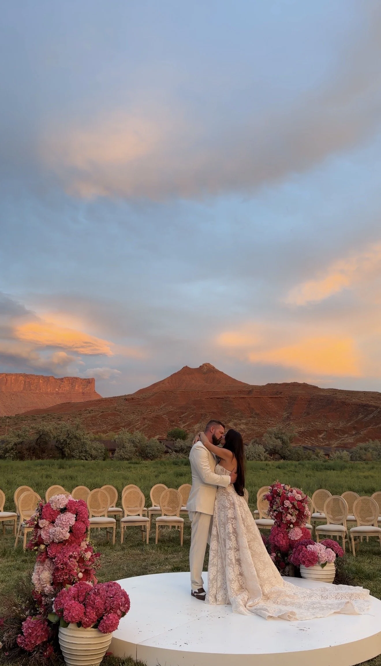 A couple is standing on a small, circular platform outdoors, embracing and kissing, with a scenic desert landscape and mountains in the background during sunset. Decorative pink and purple floral arrangements surround the platform, and empty chairs are arranged behind them for a wedding ceremony.