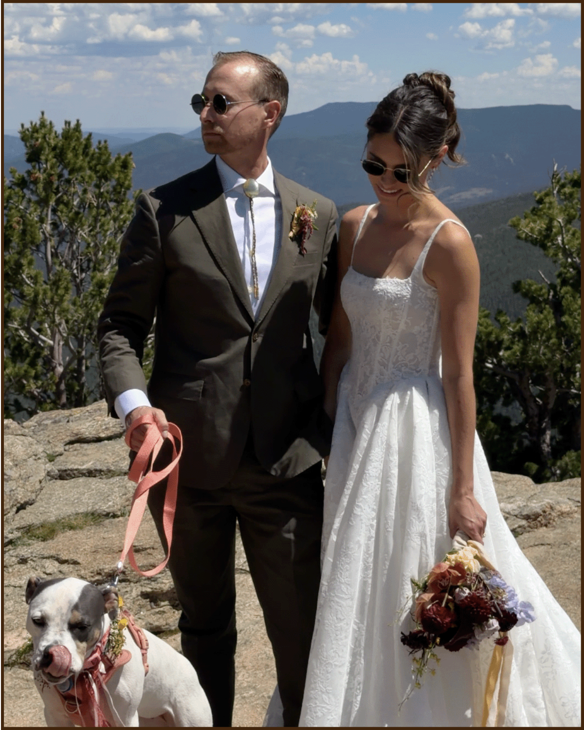 A man in a suit and sunglasses holding a leash with a dog, and a woman in a white wedding dress holding a bouquet with flowers, standing outdoors on rocky terrain with trees and mountains in the background.