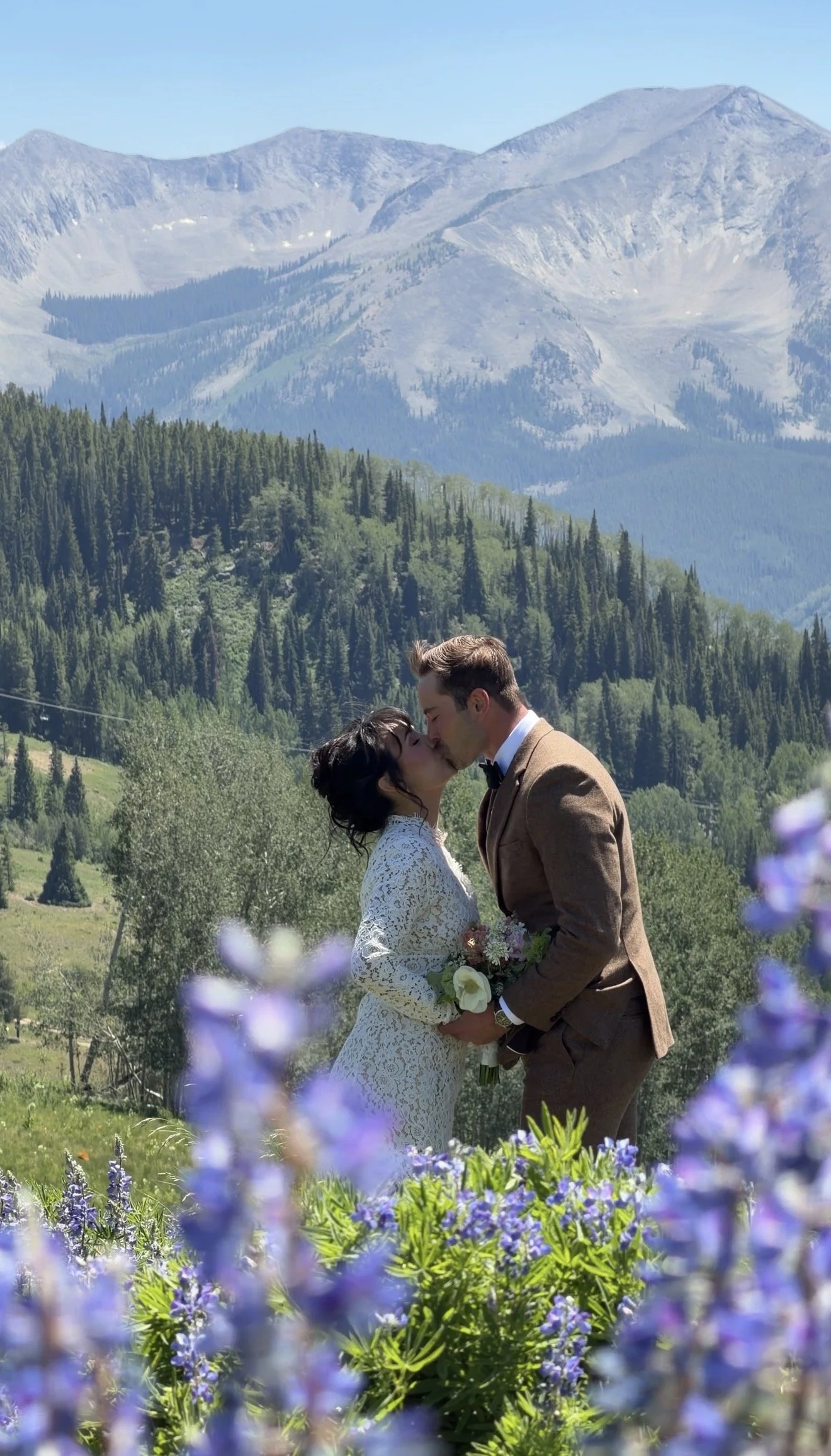 A couple in wedding attire sharing a kiss outdoors with a mountain landscape, forested hills, and colorful flowers in the foreground.
