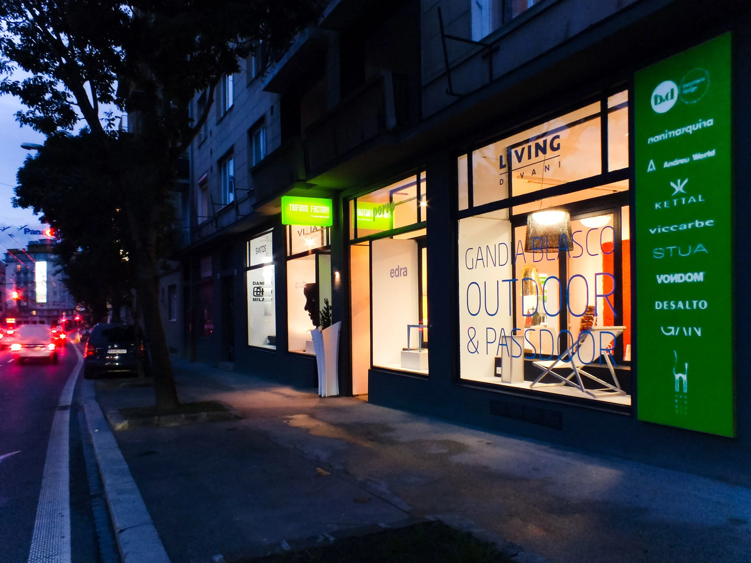 Storefront with bright green signs on a city street during early evening or dusk, showing reflections on the glass windows and silhouettes of trees and cars parked along the curb.