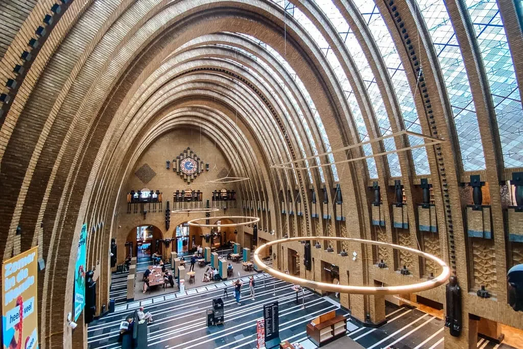 Interior of a large train station with arched brick walls, high glass ceiling, and modern circular light fixtures, with people walking and sitting inside.