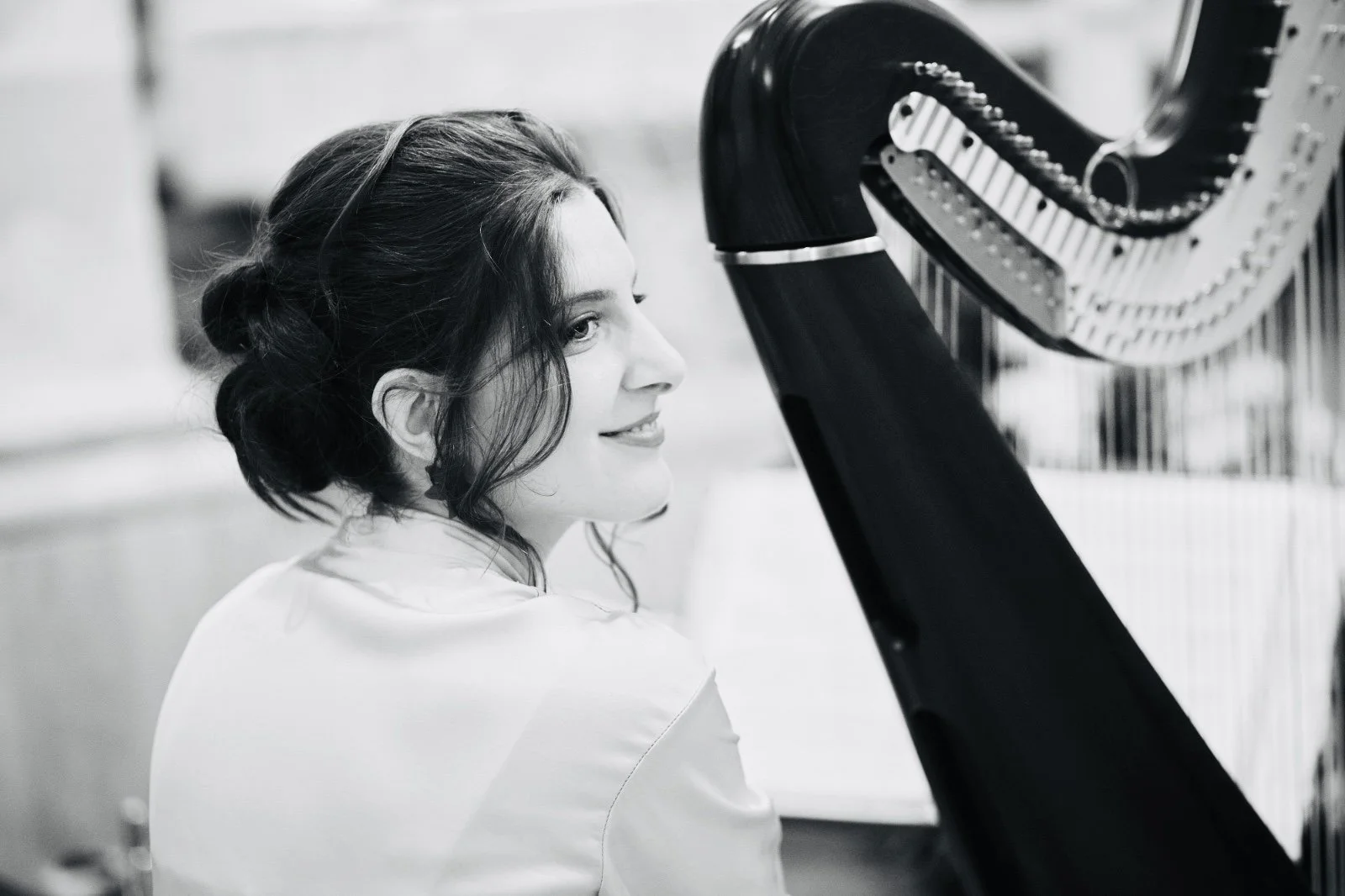 Una donna con capelli scuri e riccioli giocati, sorridente, seduta accanto a un pianoforte a coda nero in un ambiente interno.