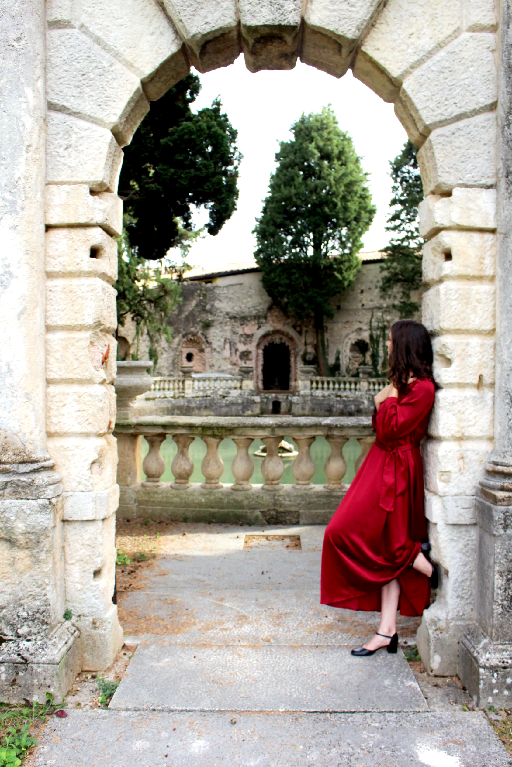 Una donna in un vestito rosso scuro sta appoggiata a un'arcata di pietra, con un cortile antico in background, alberi e un muro di pietra.