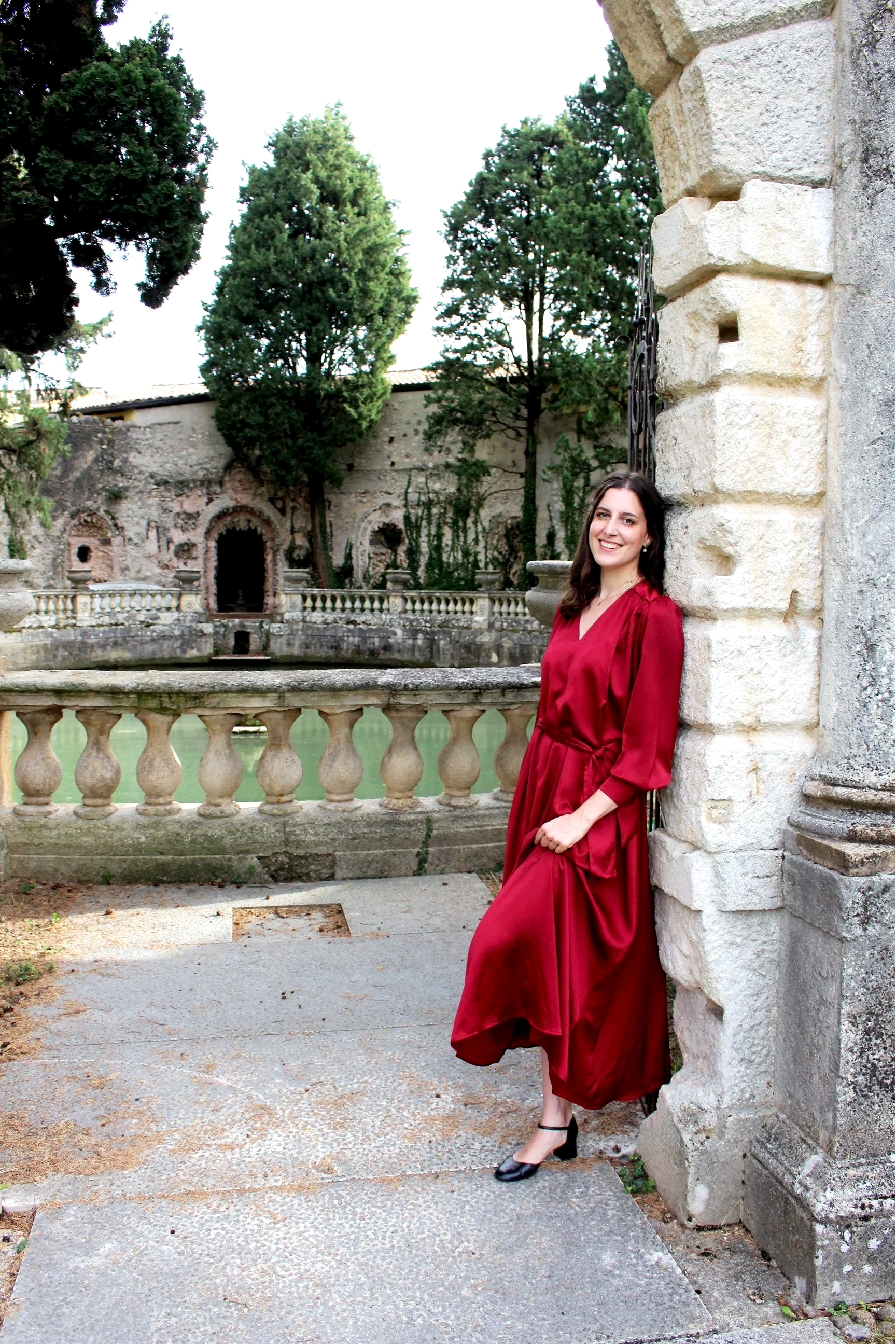 Una donna sorridente in abito rosso scuro si appoggia a un muretto in pietra di fronte a una fontana antica circondata da alberi verdi
