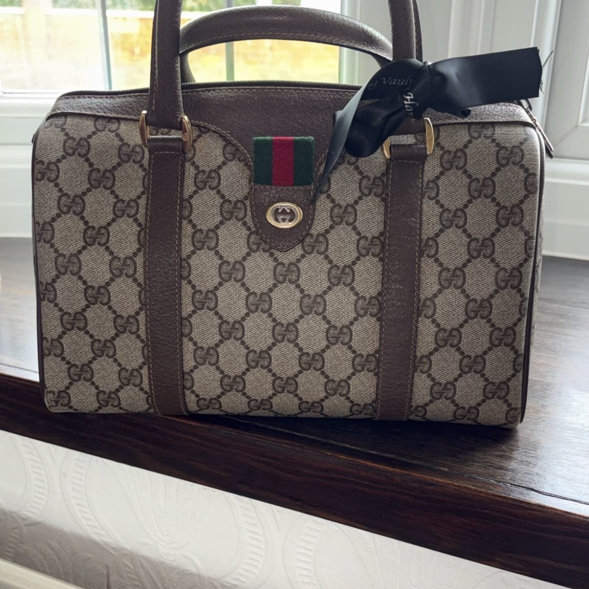A Gucci travel bag with the brand's signature monogram pattern, brown leather trim, handles, and a black bow attached to the strap, placed on a wooden surface near a window.